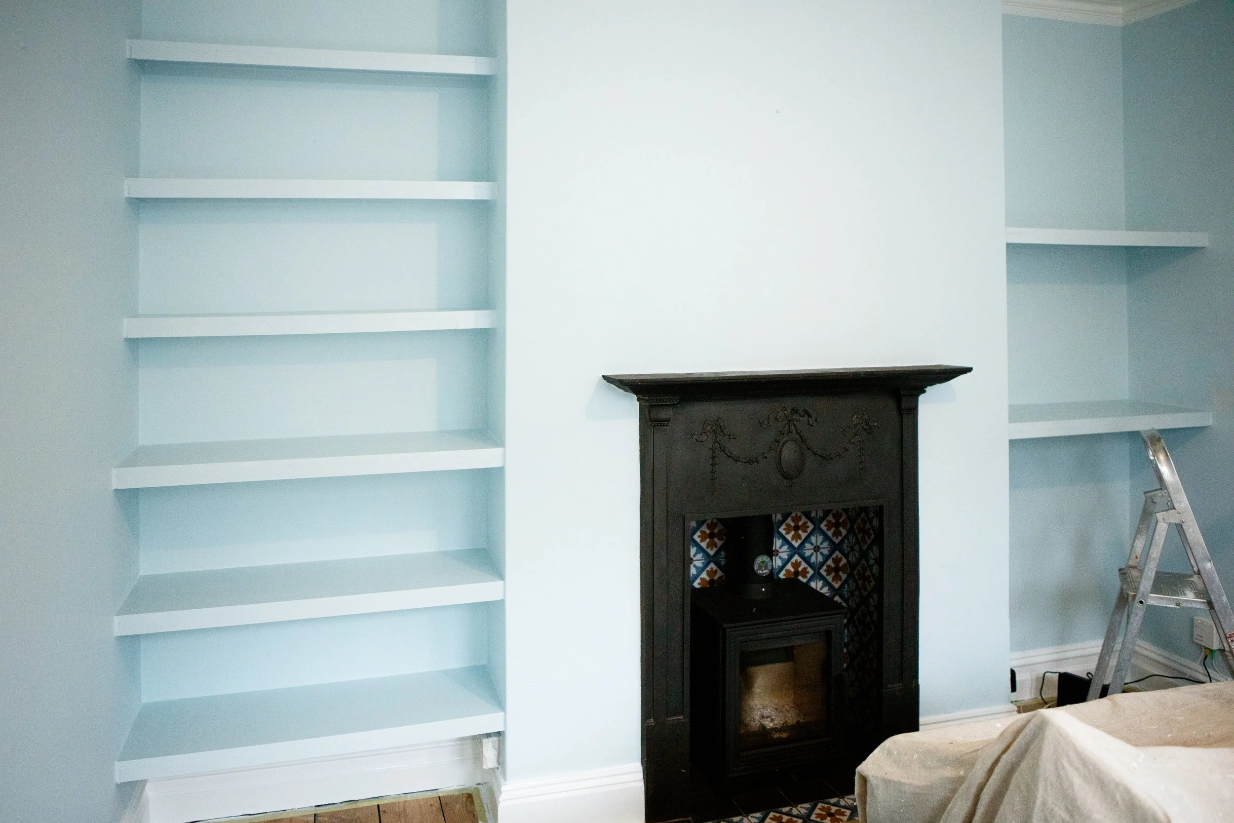 Living room with light blue walls, empty white built-in bookshelves on both sides of a black fireplace with decorative tiles, and a ladder leaning against the right wall.