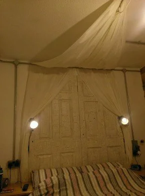 A bedroom with a bed, striped bedspread, wooden headboard, and fabric canopy draped from the ceiling with two lamps on either side.
