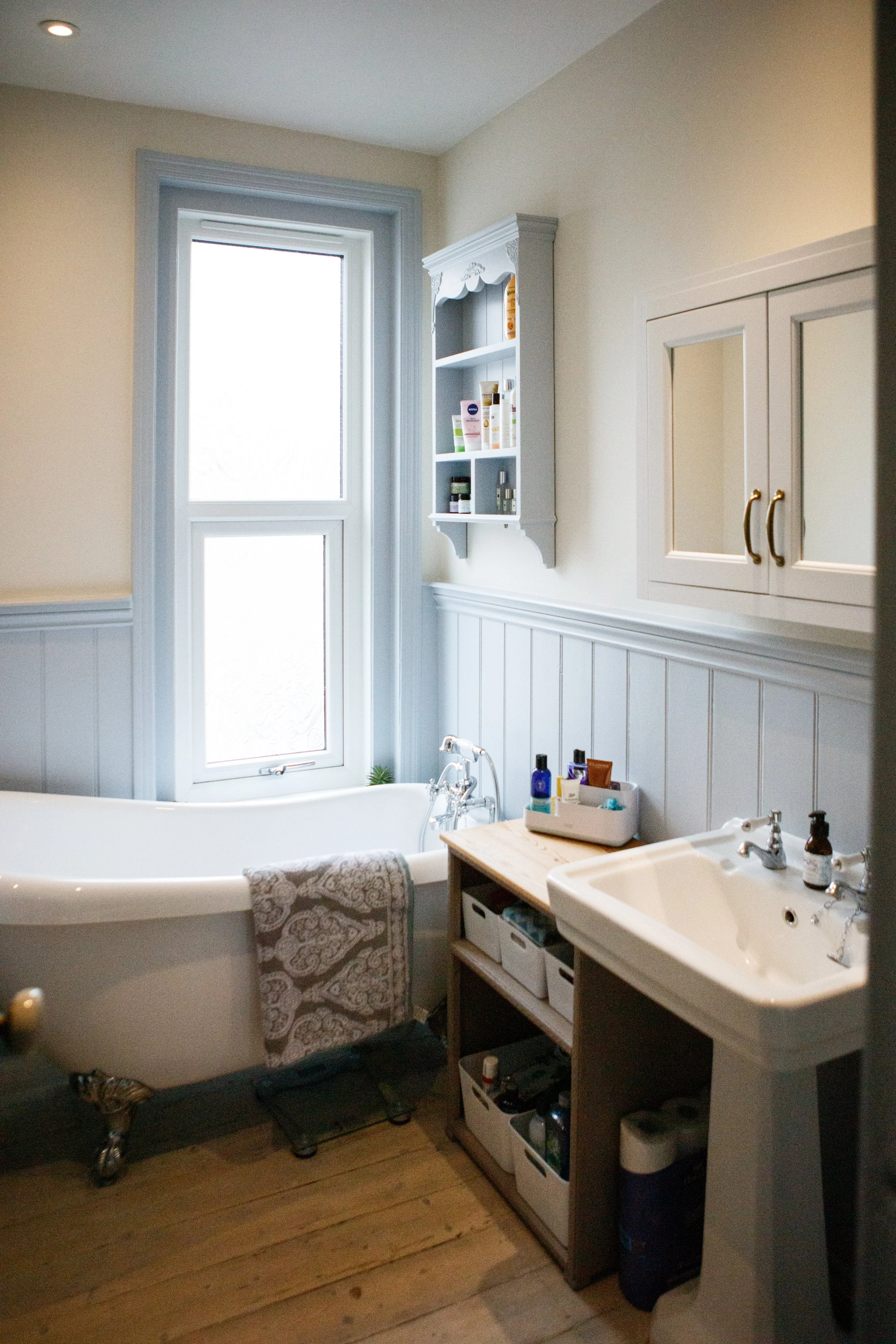 A bathroom with a clawfoot bathtub near a window, a small sink, and storage shelves with toiletries and towels.