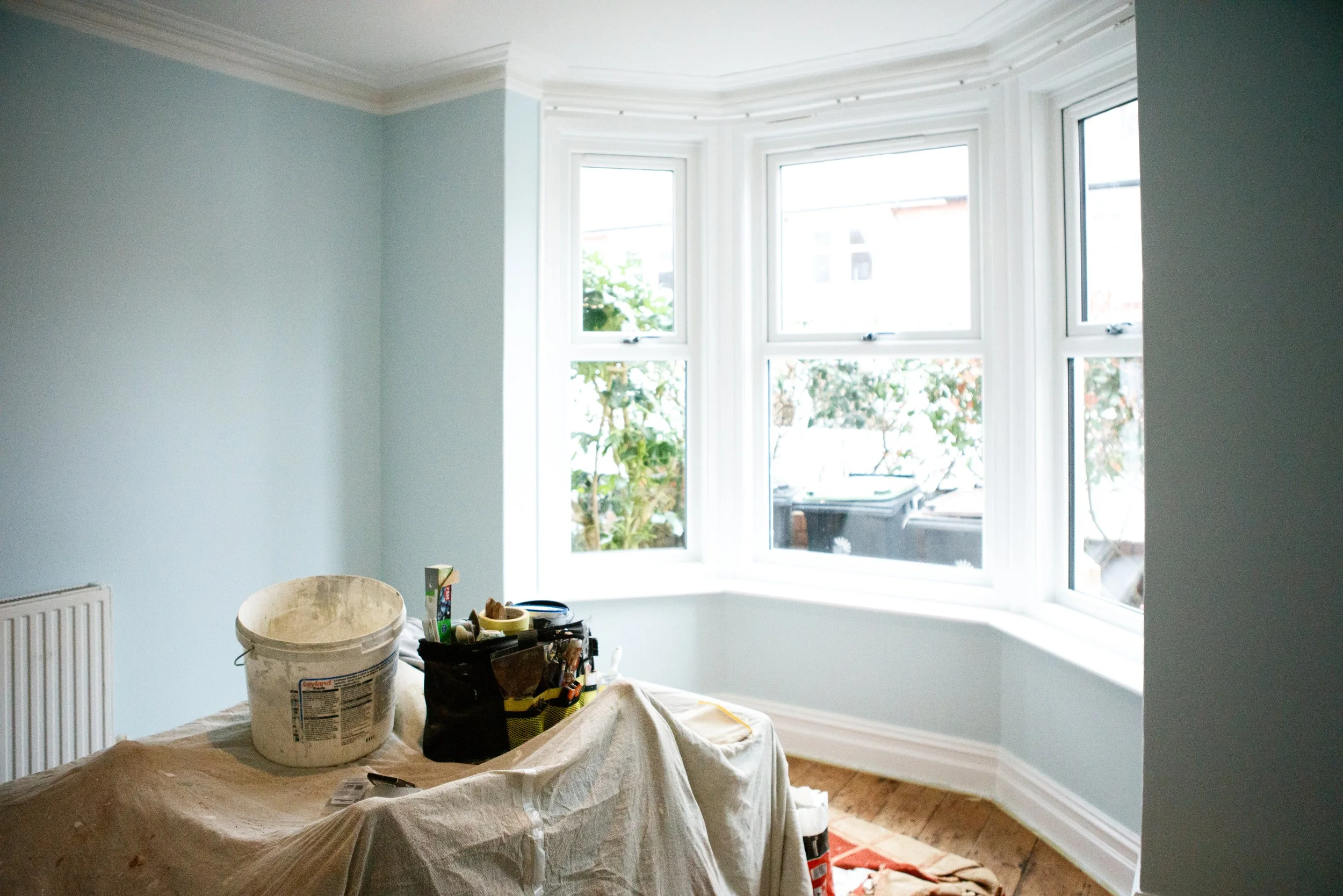 Room with light blue walls, bay window, and wooden floor. There are construction tools and supplies on a covered table in the foreground.