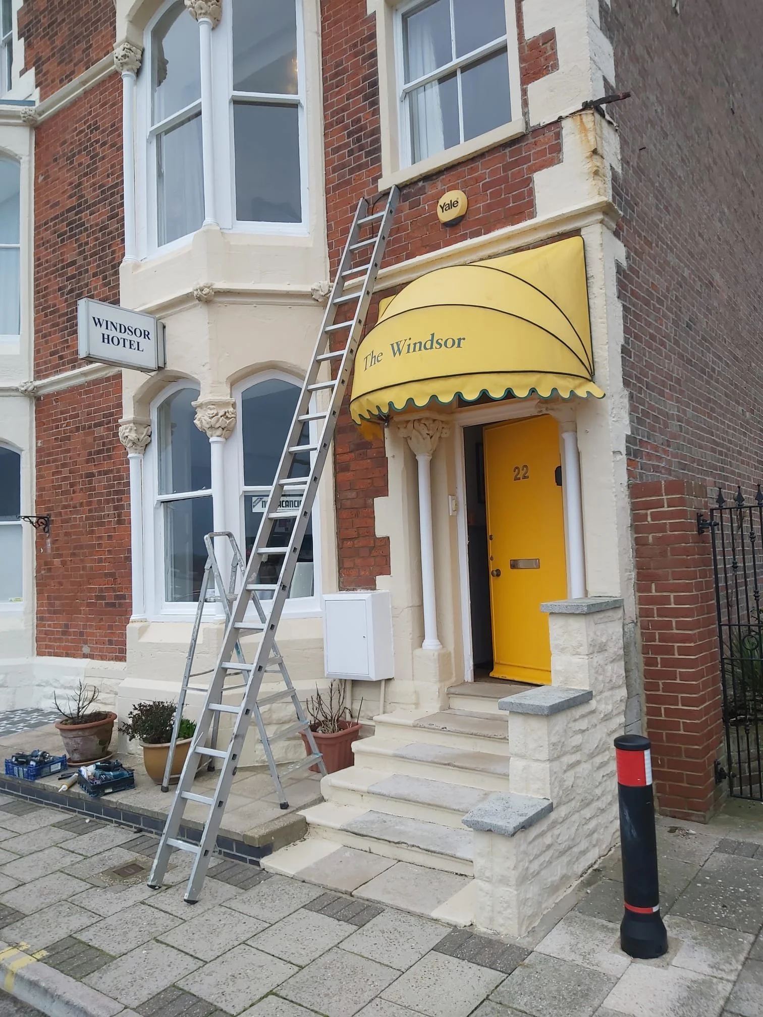 Exterior of a building with a yellow awning reading 'The Windsor' over a yellow front door, set on a step with a small black and red bollard nearby. A stepladder is leaning against the building, and a sign for 'Windsor Hotel' is mounted on the wall t