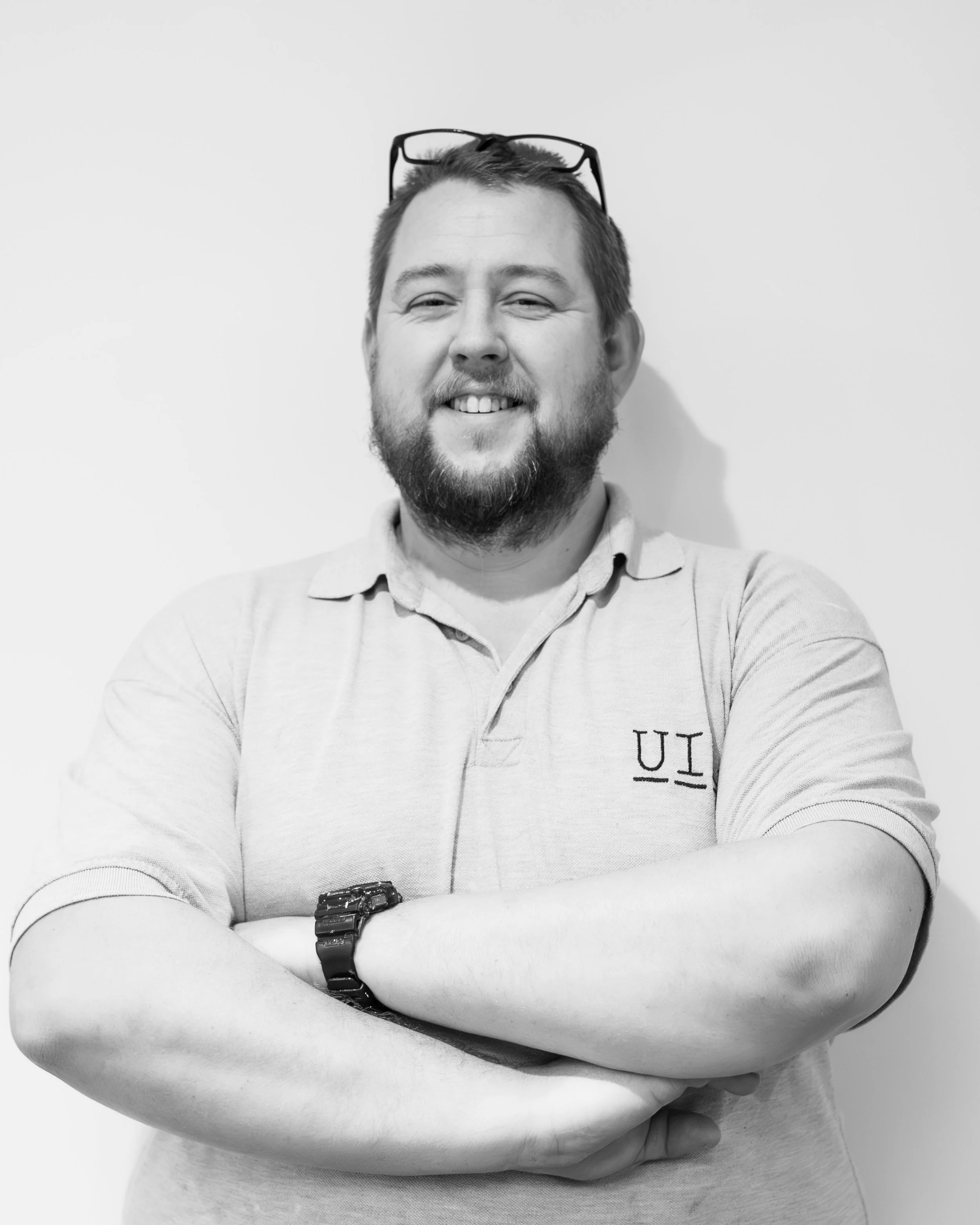 A man with shaved dark hair, a goatee beard, wearing a white polo shirt, Standing leaning against a wall smiling..
