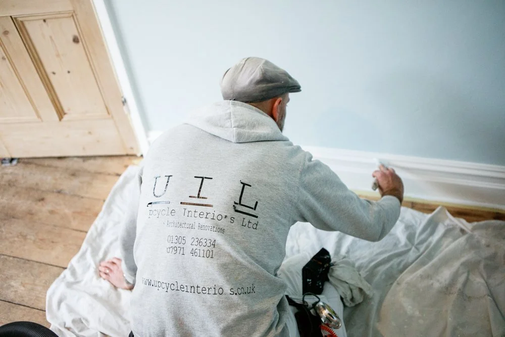 A man installing baseboard molding in a room, wearing a gray hoodie with company branding, sitting on the floor with tools around him.