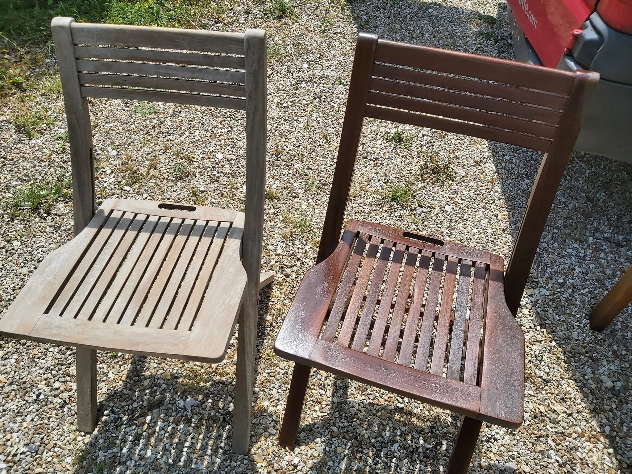 Two wooden chairs, one light gray and one dark brown, are placed outdoors on gravel ground, with a red vehicle partially visible on the right side of the image.