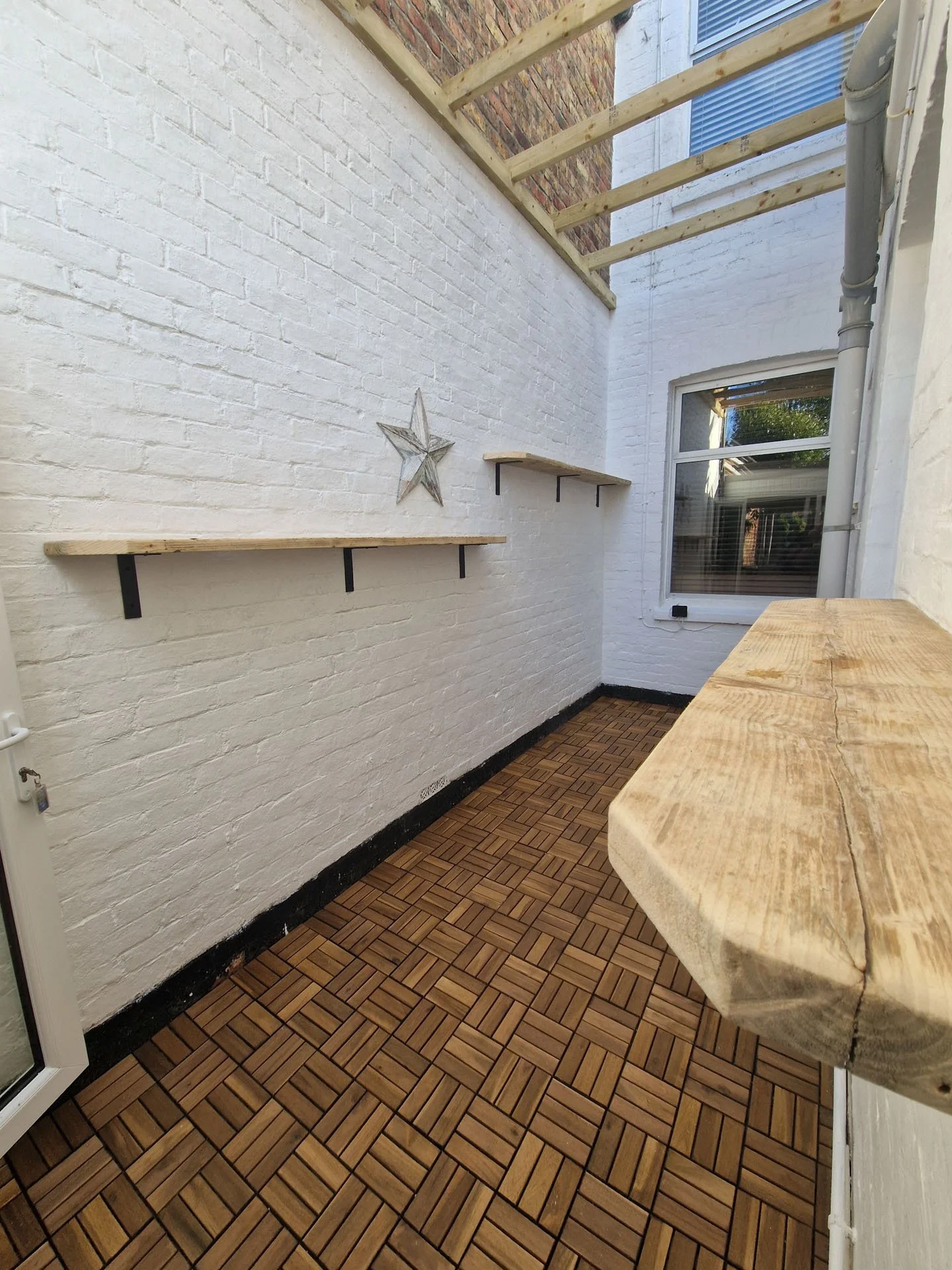 A small outdoor balcony with white brick walls, wooden shelves, and a wood-patterned tiled floor. There is a window with blinds and a star decoration on the wall.
