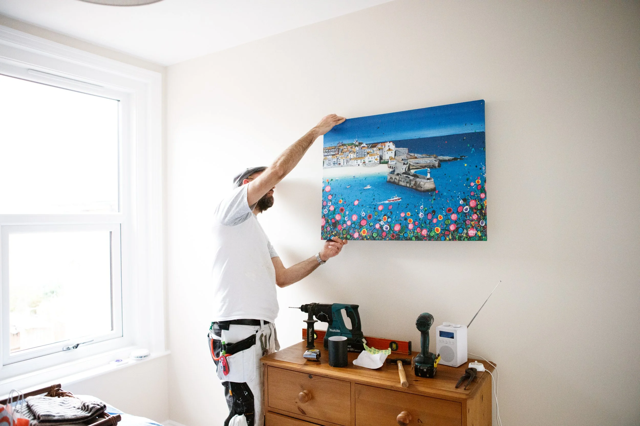A man hanging a colorful painting of a seaside town on a white wall in a bright room with a window.