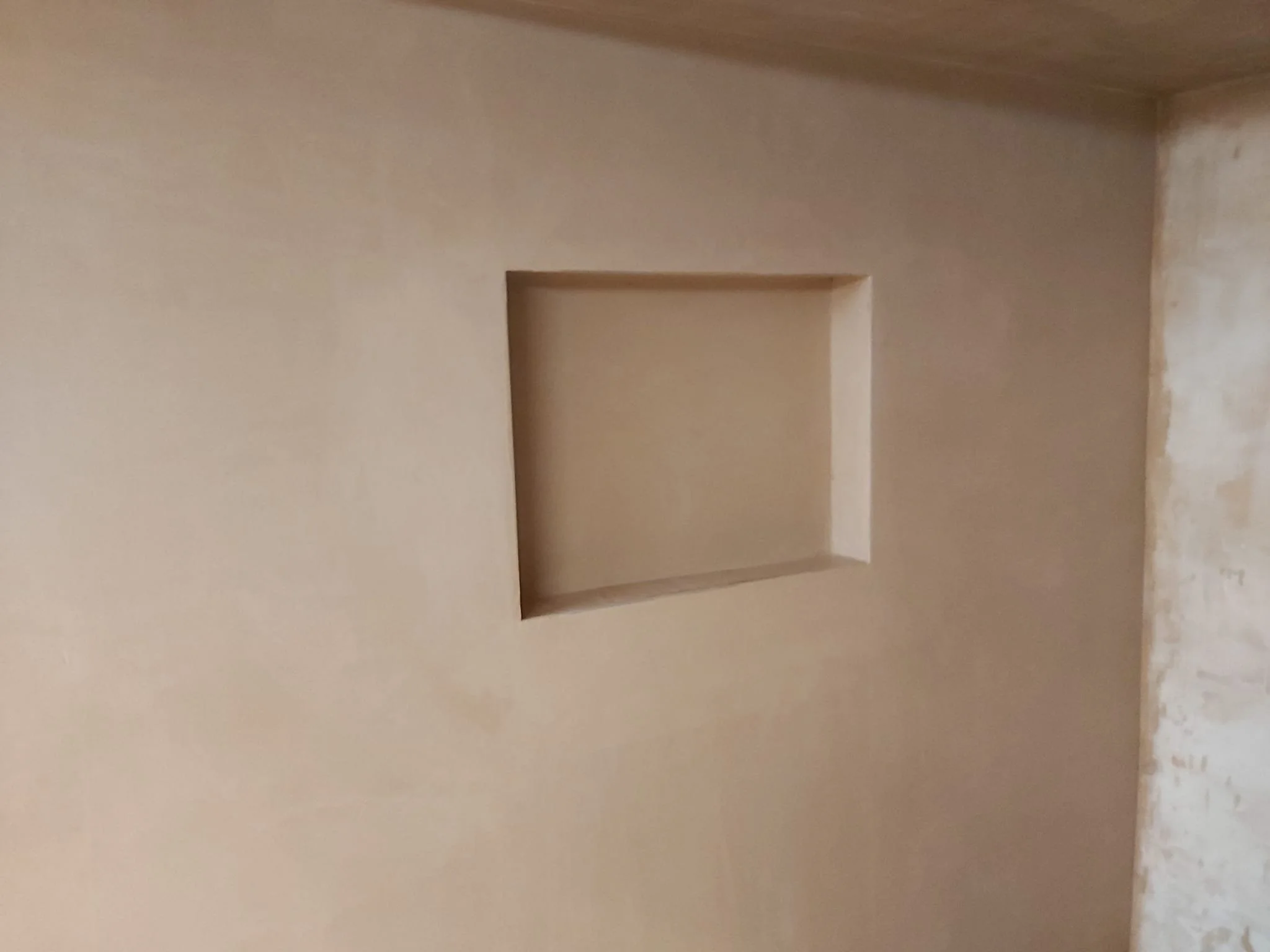 A beige wall with a square recessed shelf in the center, and an unfinished concrete floor on the right side.