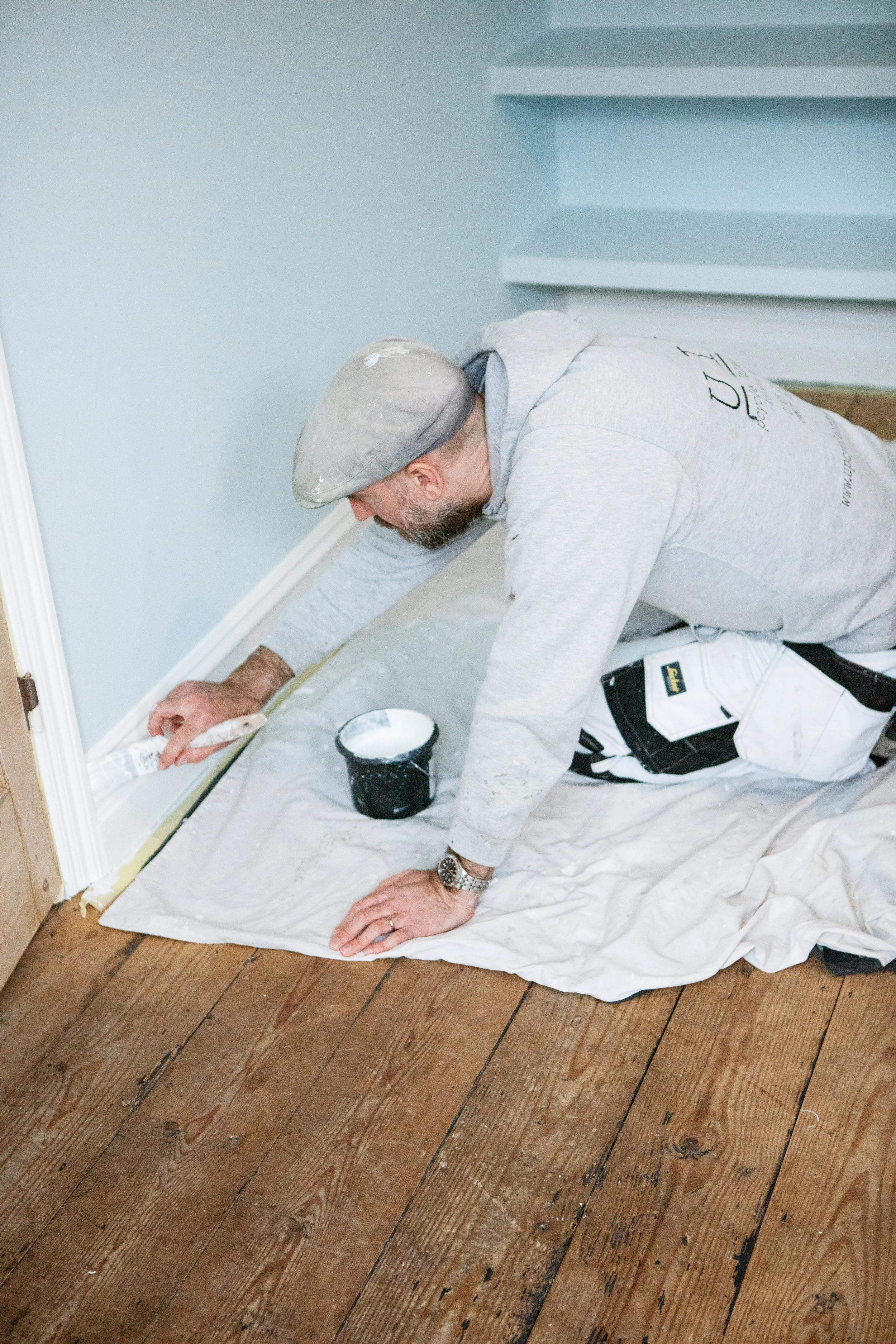 A man with a bearded face, wearing a gray cap, gray hoodie, and white pants, kneeling on the floor, painting a baseboard with white paint. He is using a small brush, and there is a black container of white paint nearby. The man is on a white drop clo