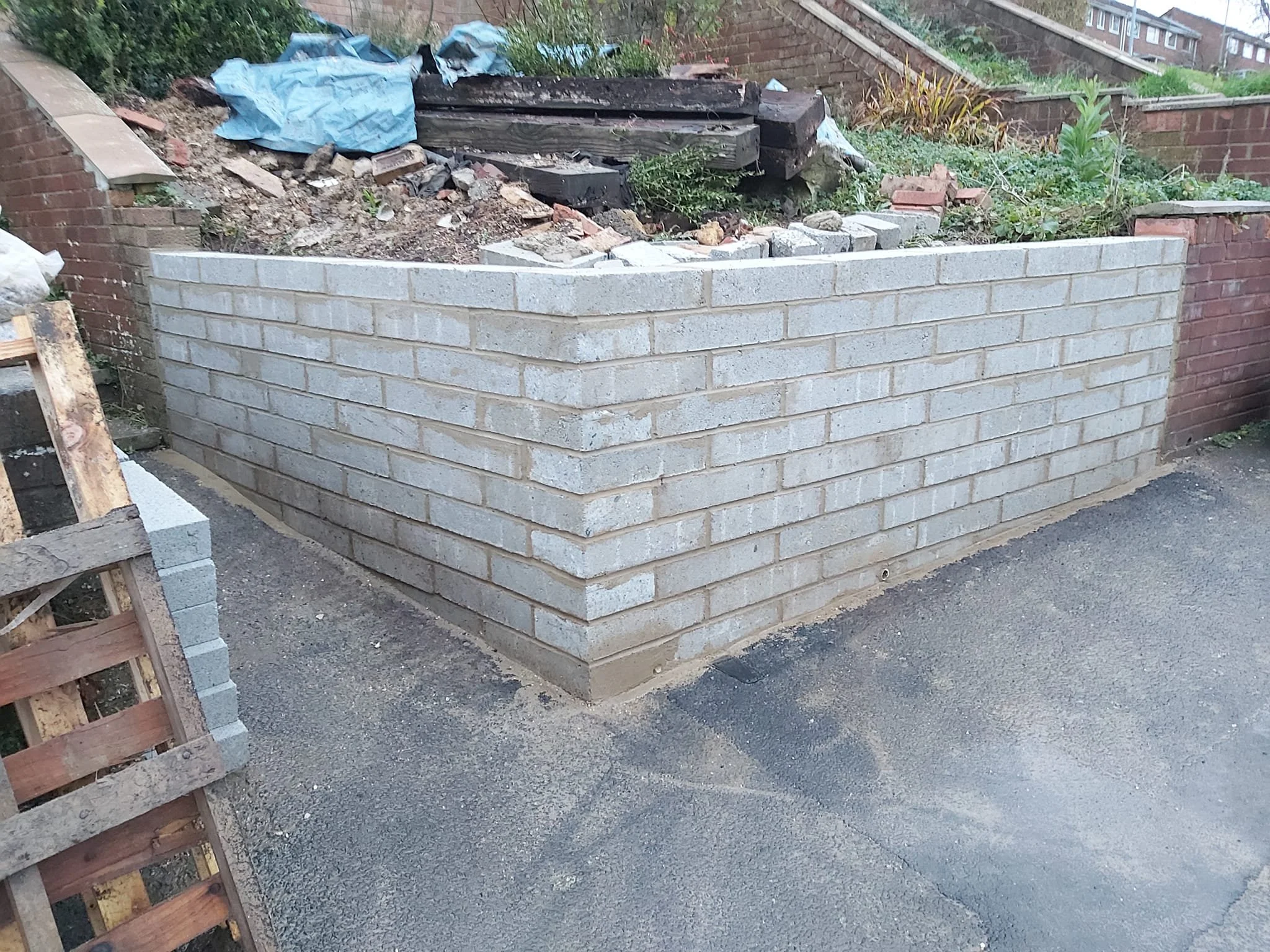 A newly built grey brick wall on a paved sidewalk, with a garden area behind it containing soil, plants, and some construction materials.