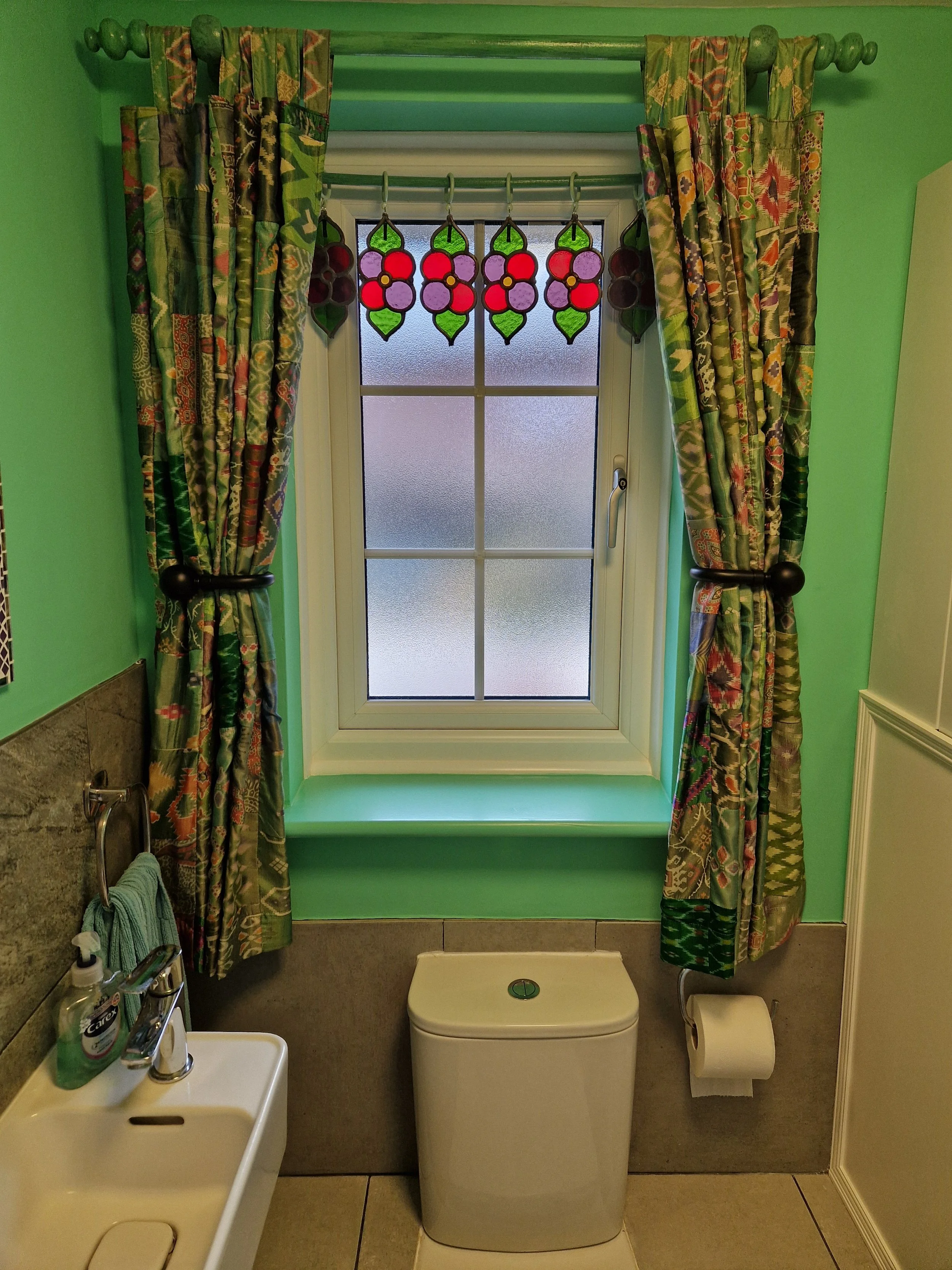 Bathroom with green walls, a window with stained glass fruit design, floral curtains, a small sink with hand soap, a toilet, and a toilet paper roll.