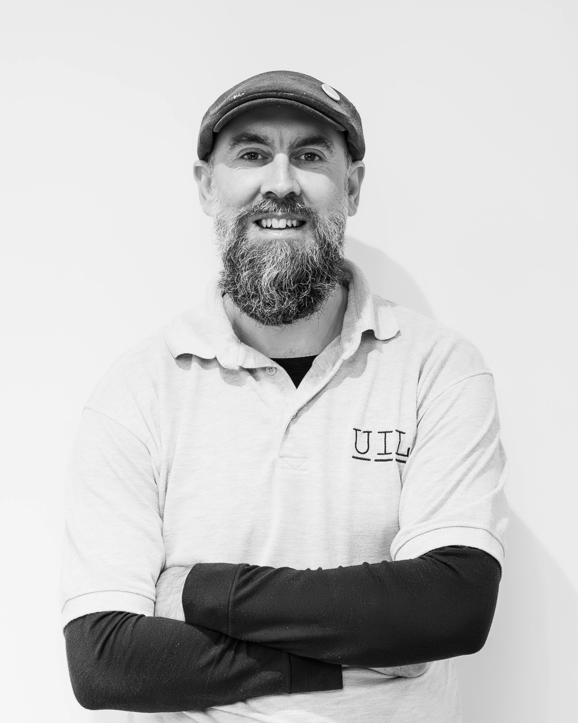A man with a grey flat cap, smiling at the camera. He is wearing a white shirt and black trousers