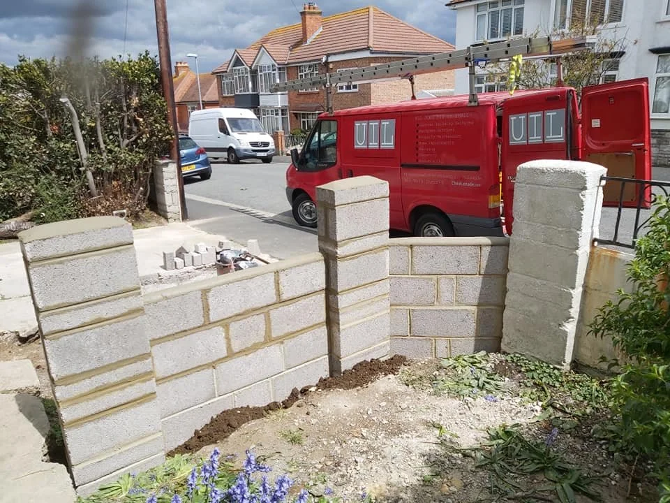 Construction of a small brick wall in a residential area on a street with parked cars, including a red utility van and a white van, with houses and bushes in the background.