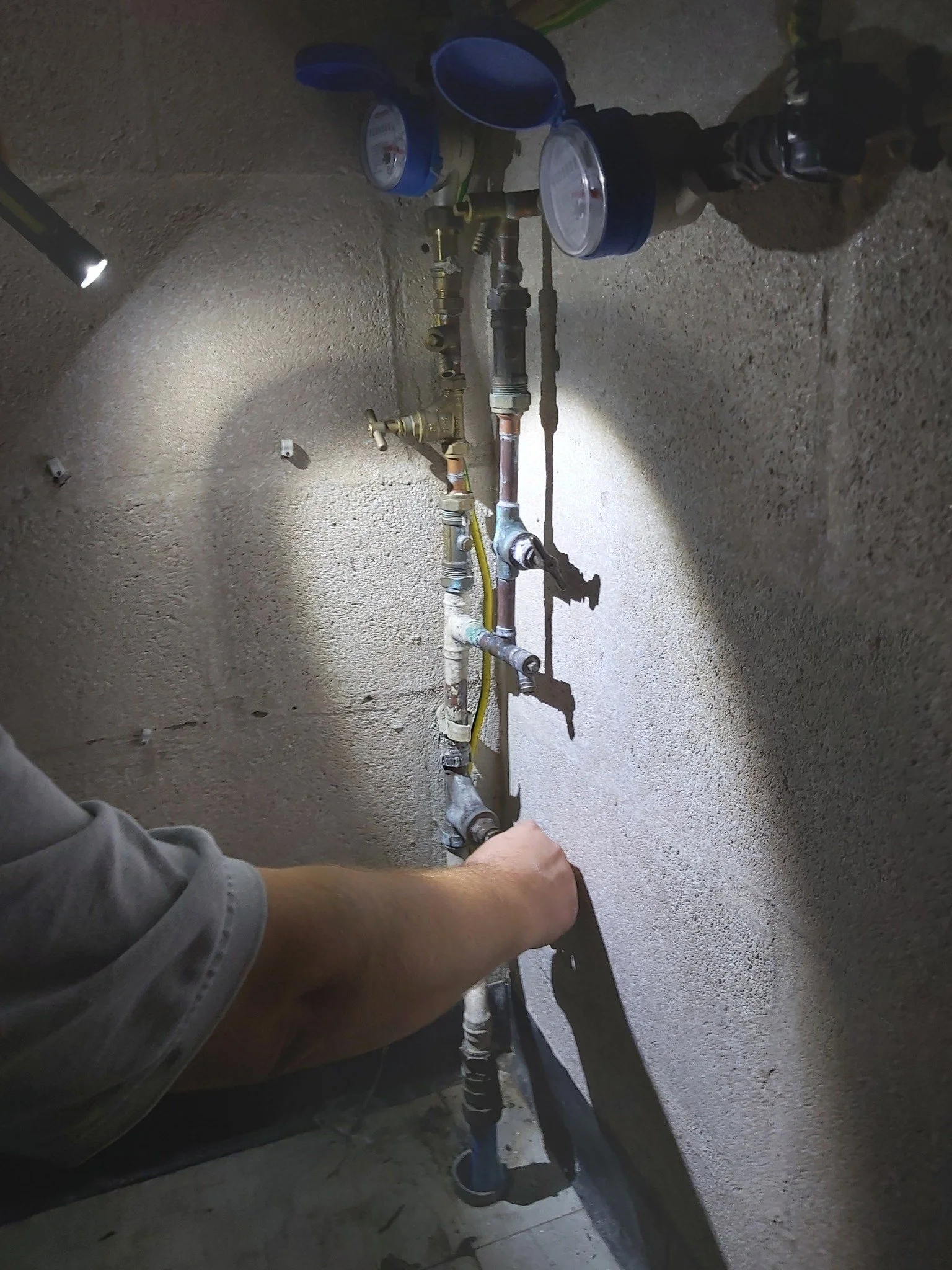 Person working on plumbing pipes in a wall with a hand holding a wrench, with tools and gauges on the floor nearby.