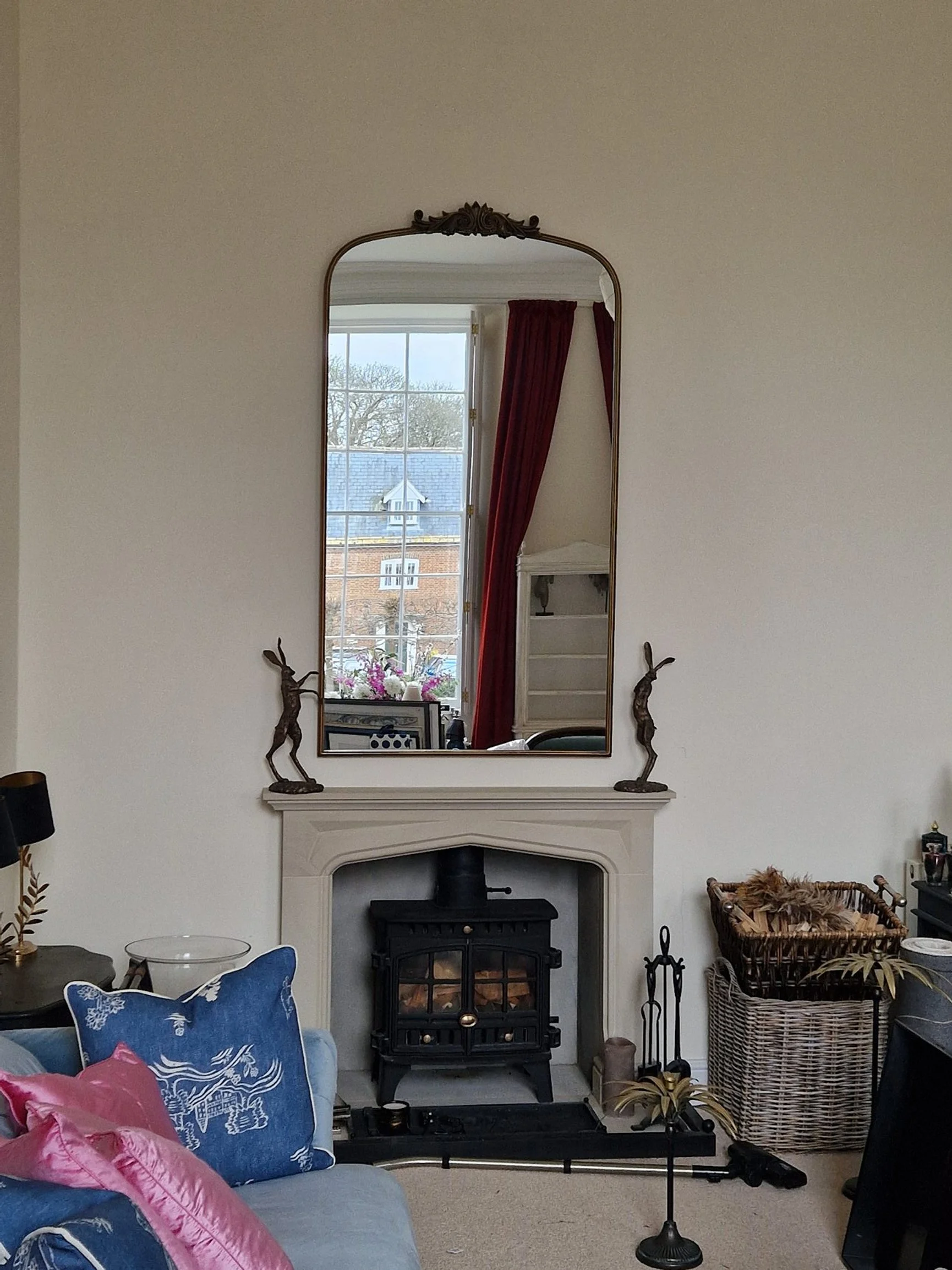 Living room fireplace with a large mirror above it, flanked by two decorative sculptures of reindeer. In the reflection of the mirror, a window with red curtains and a view of a tree and neighboring rooftops can be seen. The fireplace has a wood stov