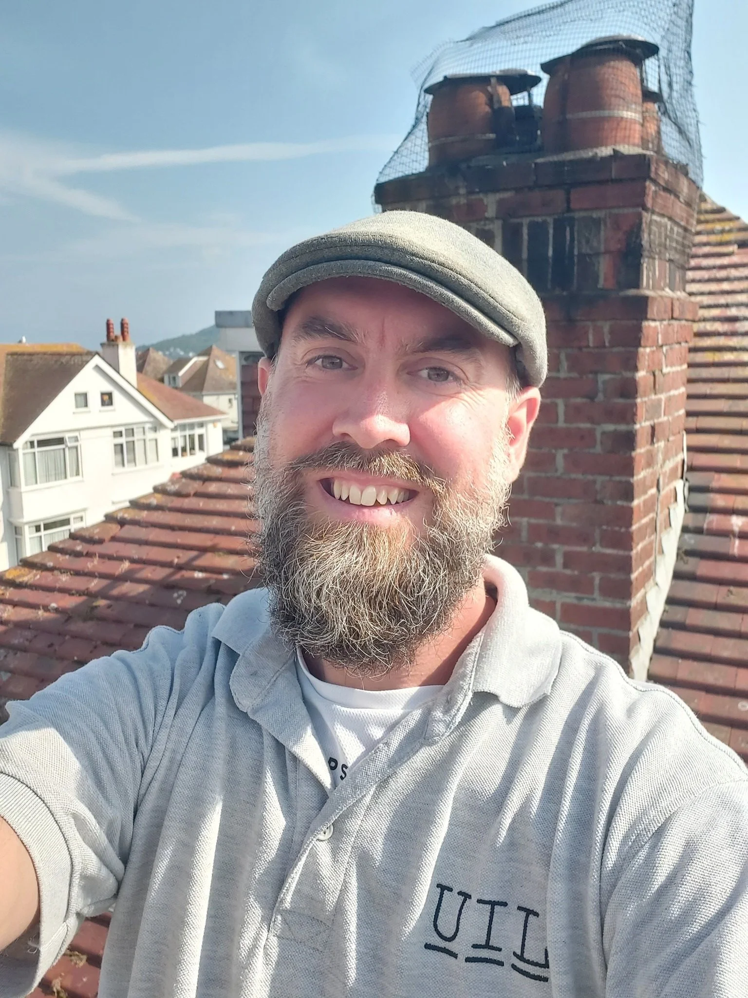 A smiling man with a beard and mustache wearing a gray cap and a light gray polo shirt taking a selfie on a rooftop with chimneys and neighboring houses in the background.
