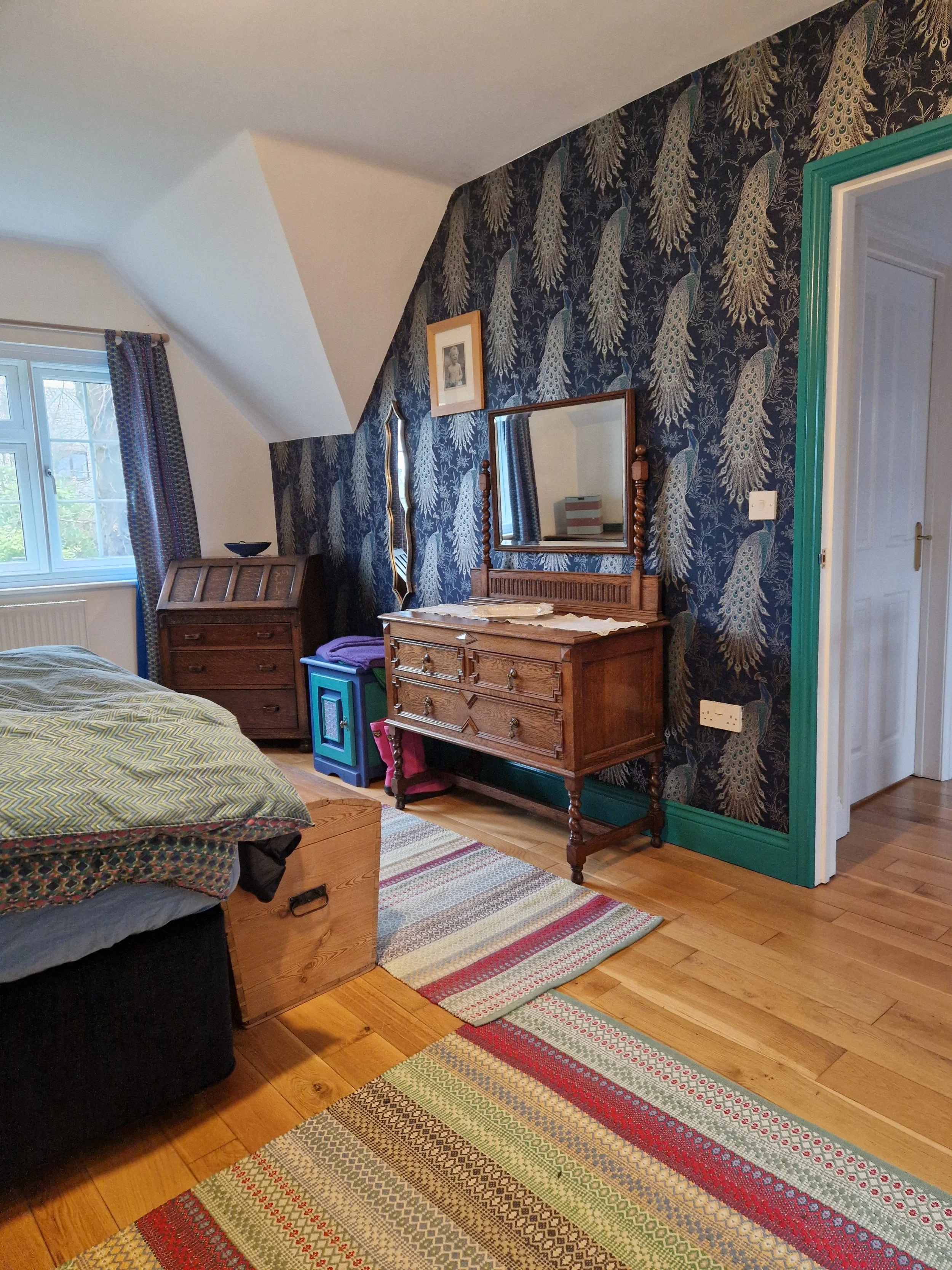 A bedroom with a wooden floor and colorful striped rugs. A bed with patterned bedding is partially visible on the left. An antique wooden dresser with a mirror is against a dark patterned wallpapered wall. A window with patterned curtains is on the f