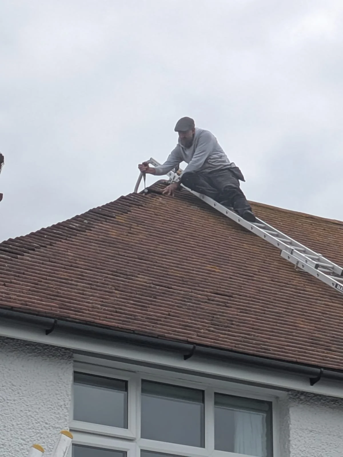 A man is working on the roof of a house, lying on a ladder while cleaning or repairing the roof tiles.