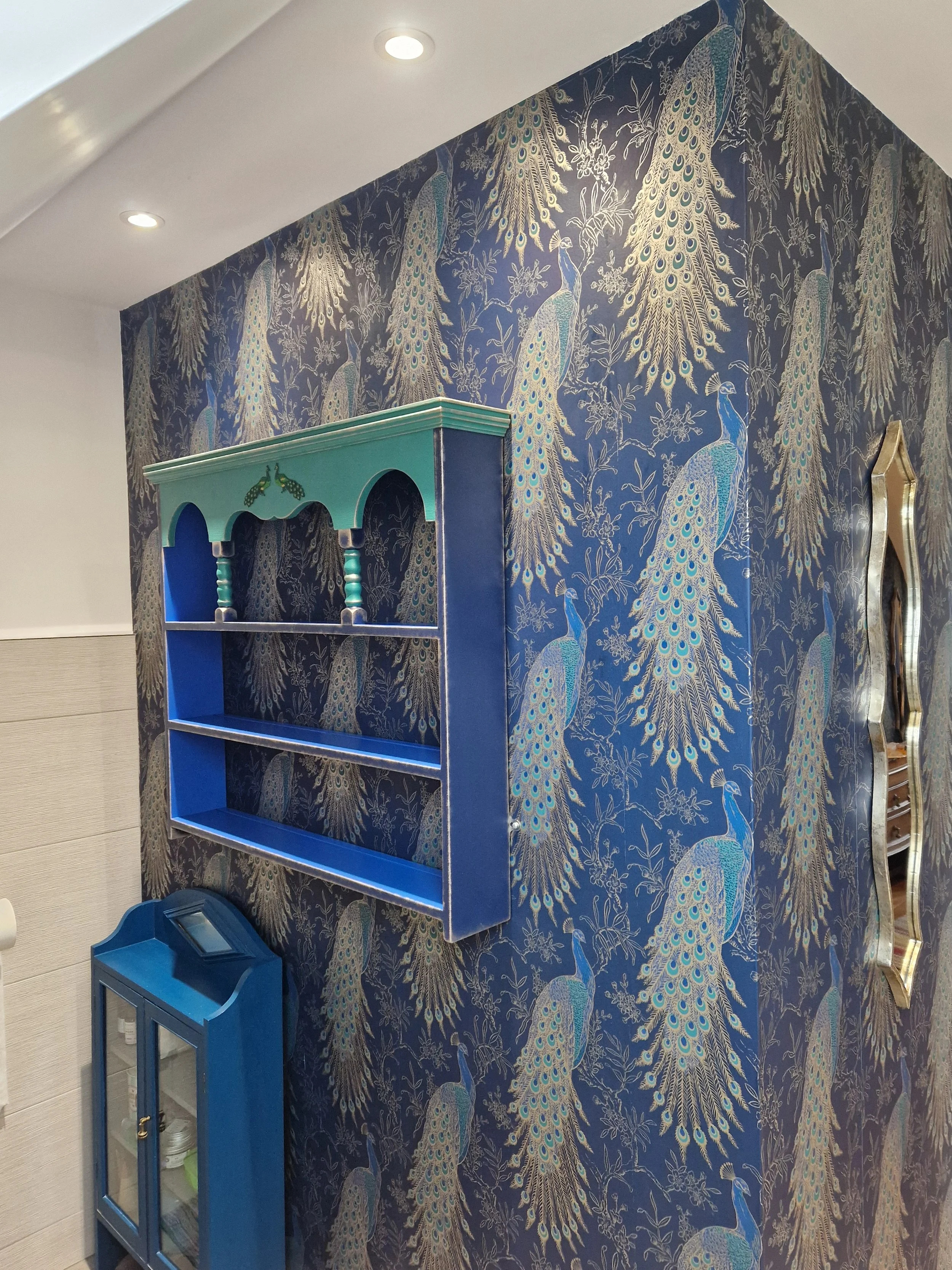 Interior wall decorated with blue and gold peacock patterned wallpaper, featuring a blue wooden wall shelf with arched top panels and a small cabinet with glass doors at the bottom.
