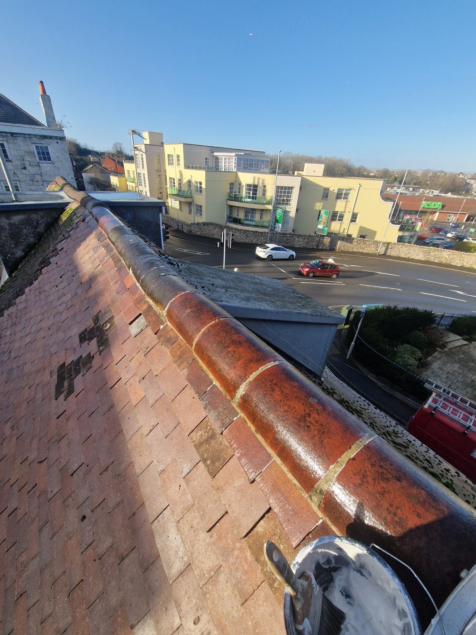 View from a rooftop showing roof tiles, a chimney, and a street with cars, buildings, and a shopping center in the background on a clear day.