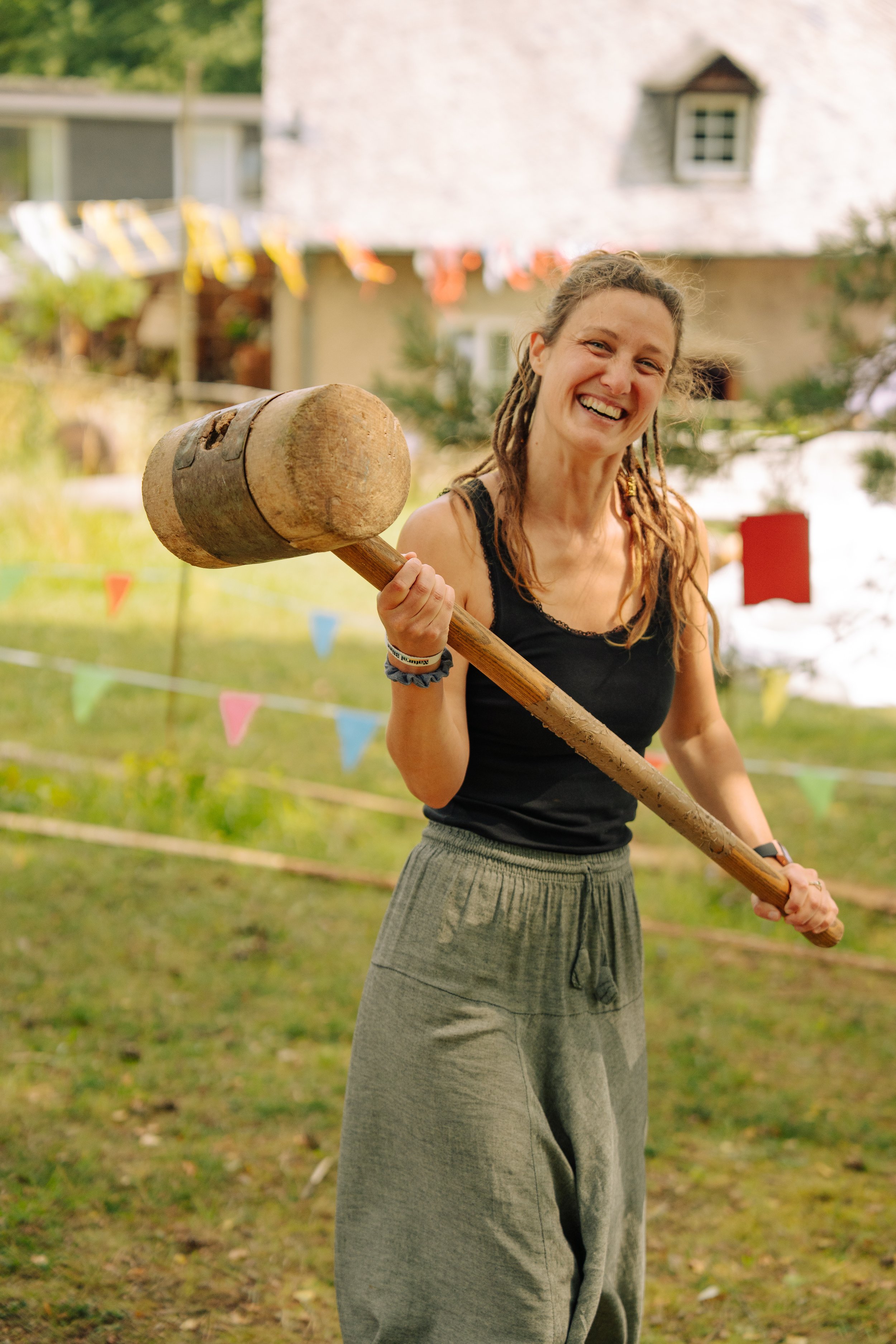 Eine lachende Frau mit Dreadlocks hält einen Holzhammer in der Hand bei einem freundlichen Outdoor-Event, im Hintergrund festliche Fahnen und ein Gebäck.