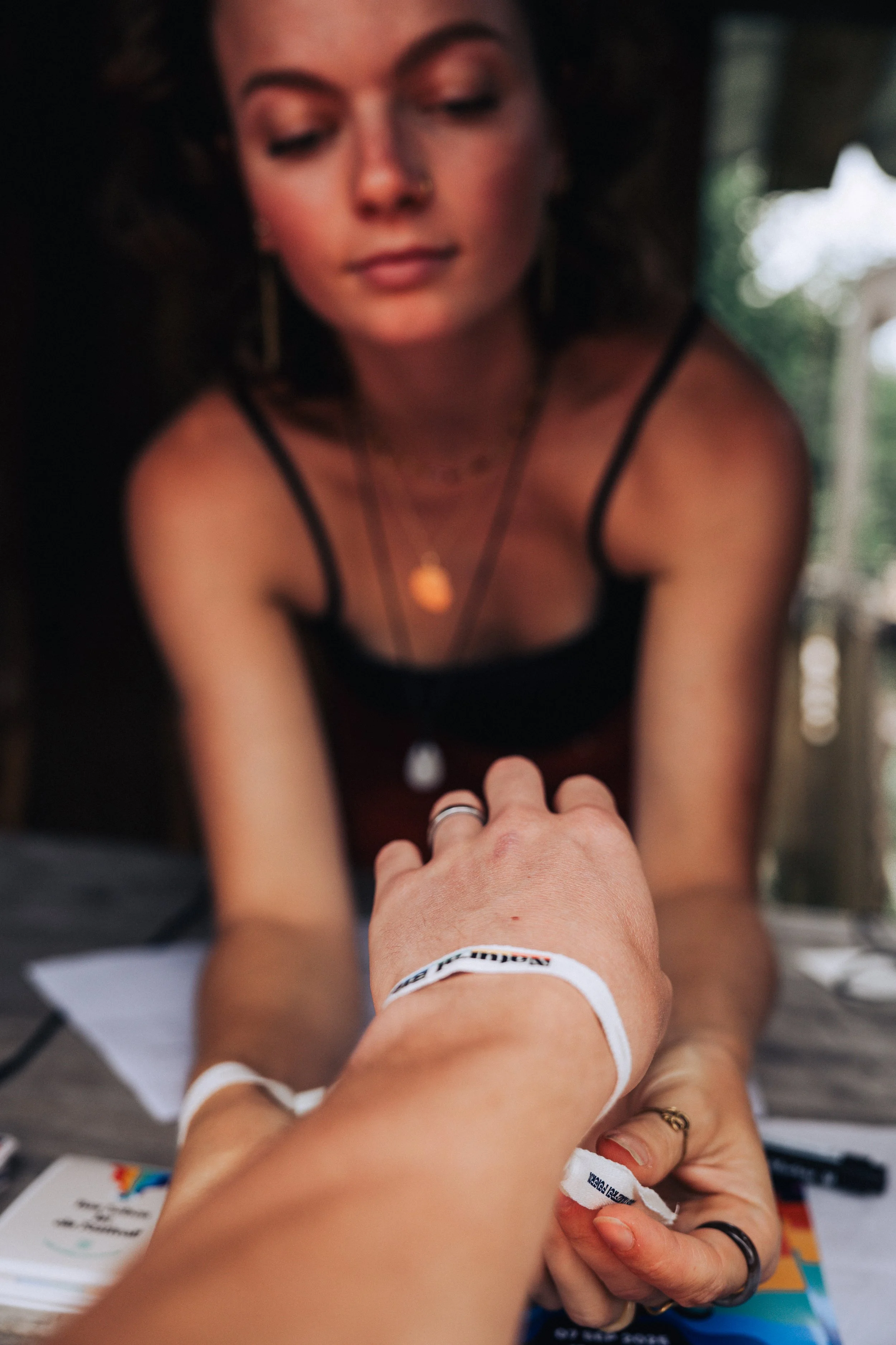 Eine Frau mit dunklen, lockigen Haaren, die eine Spitze nach vorne beugt, reicht die Hand einer anderen Person. Beide tragen Armbänder, und die Frau trägt Schmuck und dunkle Kleidung. Es ist ein Nahaufnahme-Foto, das eine freundliche Interaktion in einem natürlichen Umfeld zeigt.