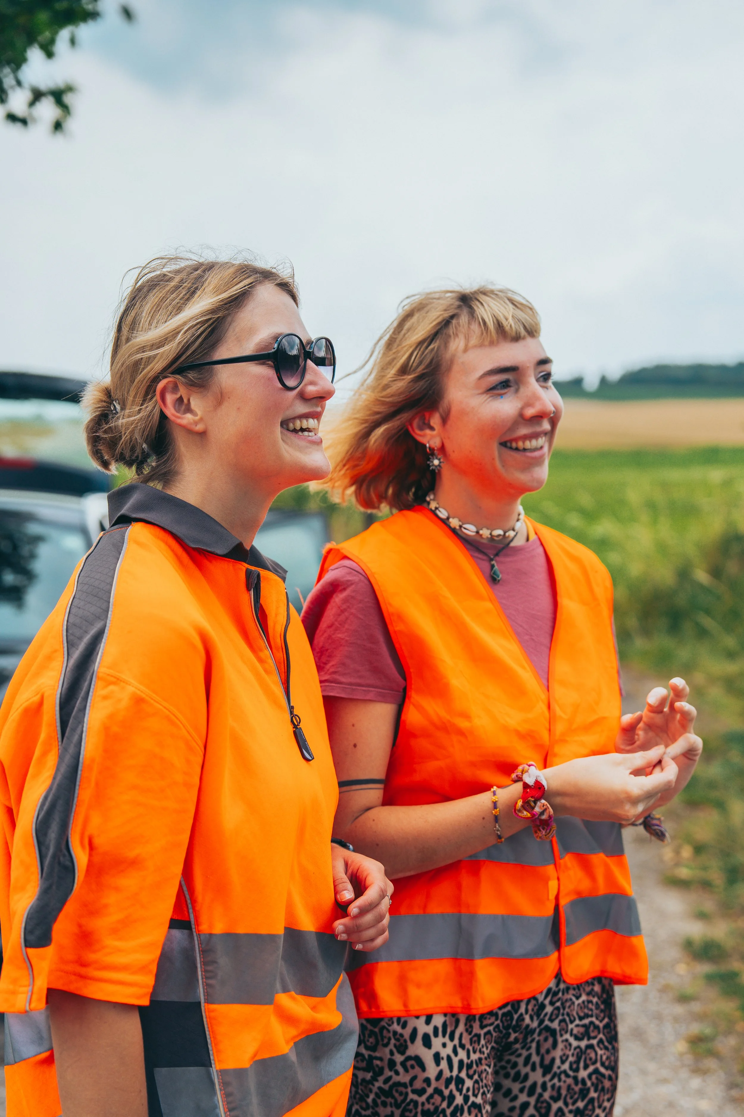 Zwei lachende Frauen in orange Warnwesten stehen draußen in einer ländlichen Gegend mit grünen Feldern und Bäumen im Hintergrund.