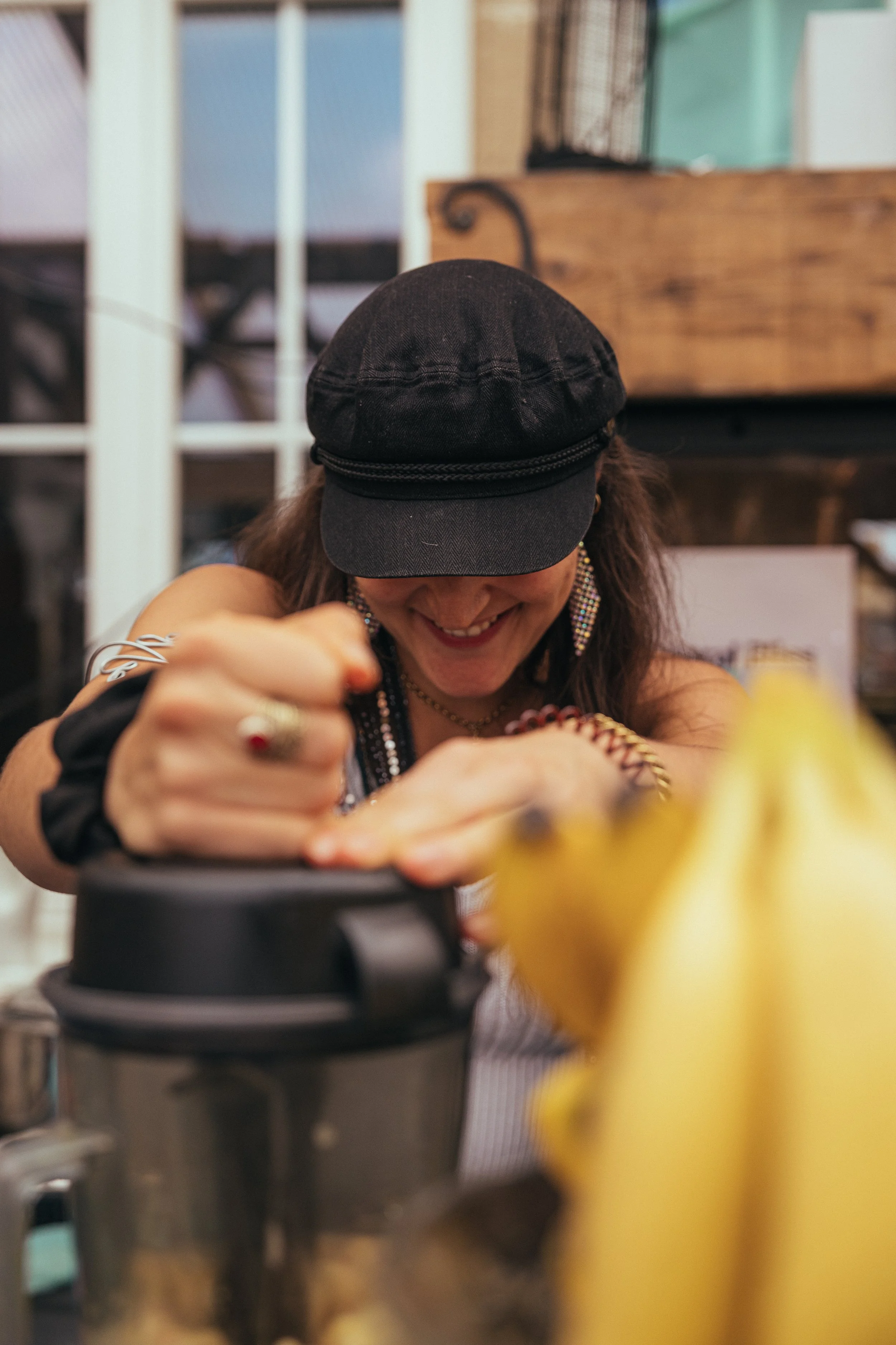 Eine lachende Frau mit dunklem Hut, die an einem Küchengerät arbeitet, im Vordergrund Bananen.