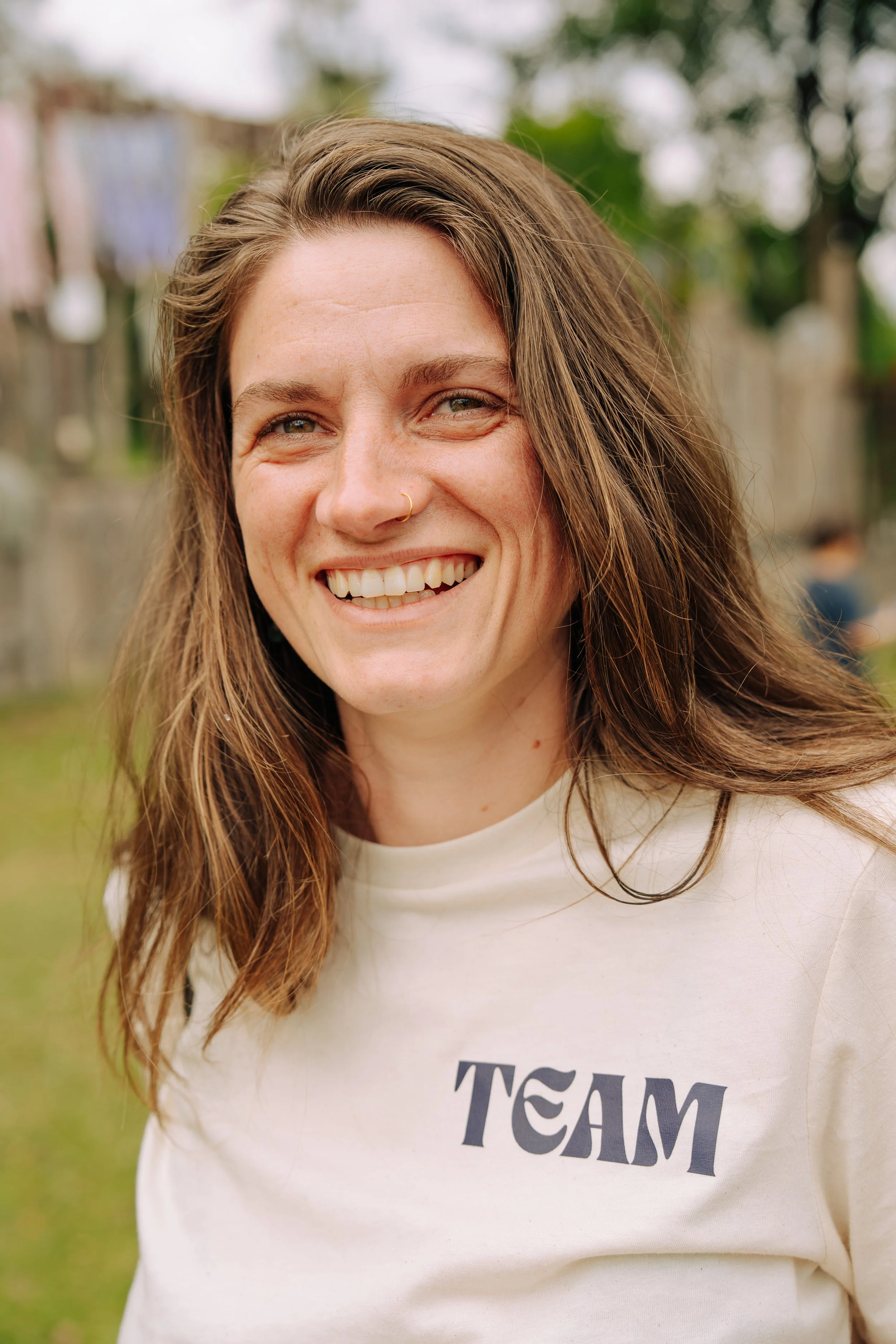 Lächelnde junge Frau mit braunen Haaren und Nasenpiercing, trägt ein weißes T-Shirt mit der Aufschrift 'TEAM', im Hintergrund eine grüne, bewaldete Umgebung.