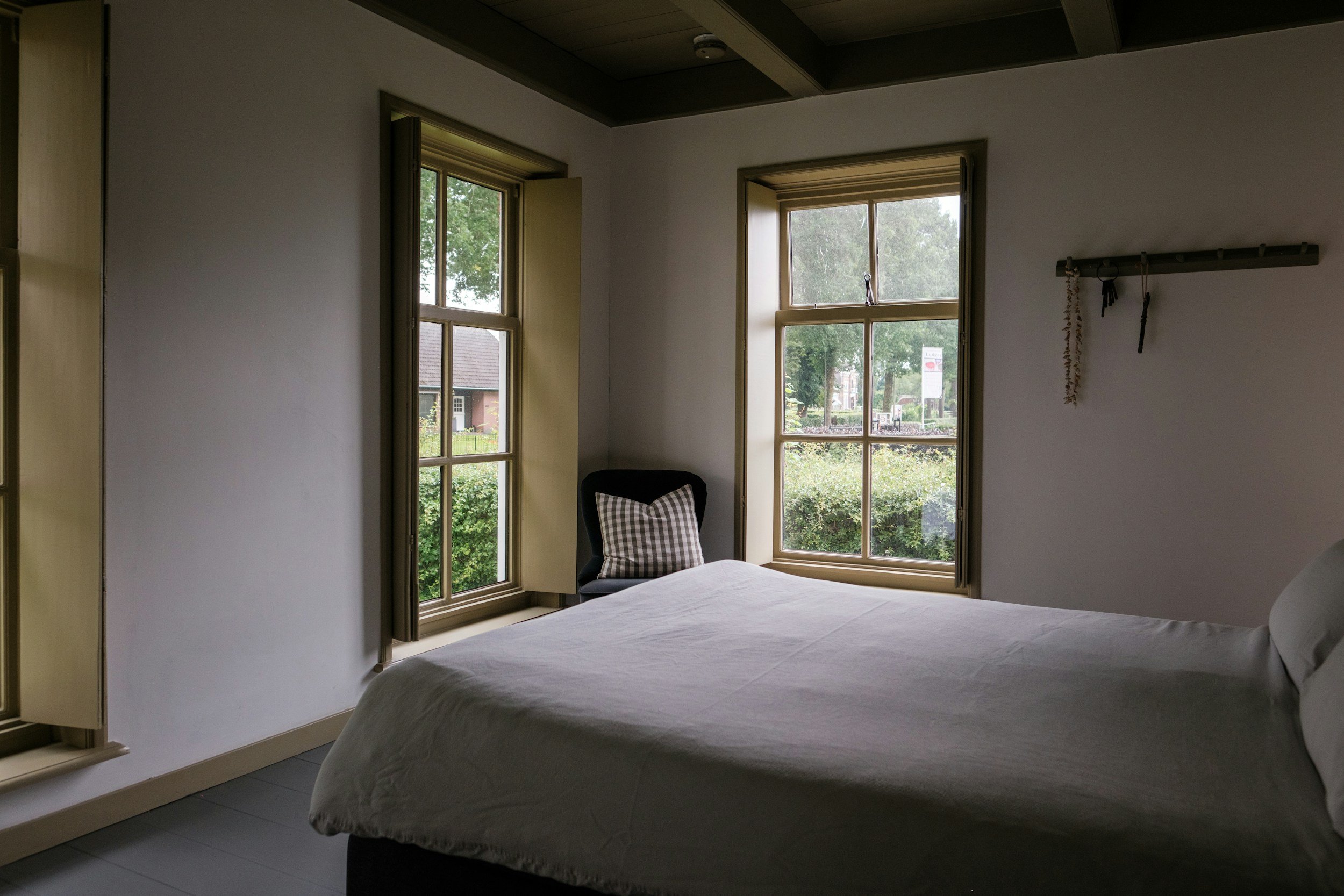 A bedroom with two large windows allowing natural light, a bed with white bedding, a black chair with a checkered pillow near the window, and wall hooks with hanging decorations.