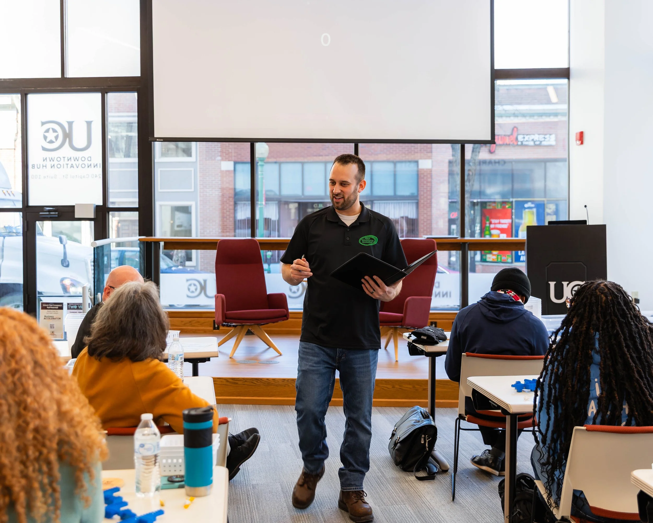 A man in a light blue shirt and dark pants pointing at a whiteboard during a presentation in a modern office setting, with a brick wall and large windows in the background.