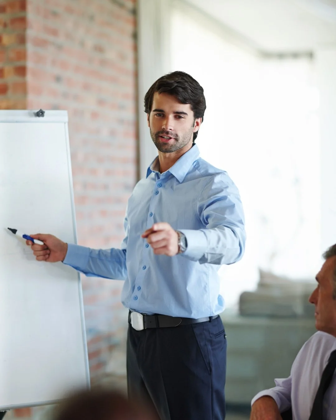 A man in a light blue shirt and dark pants pointing at a whiteboard during a presentation in a modern office setting, with a brick wall and large windows in the background.