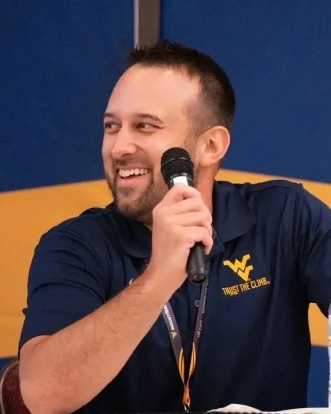 A smiling man holding a microphone during a discussion or interview, wearing a navy blue polo shirt with a yellow logo and phrase 'Trust the Climb' on it.