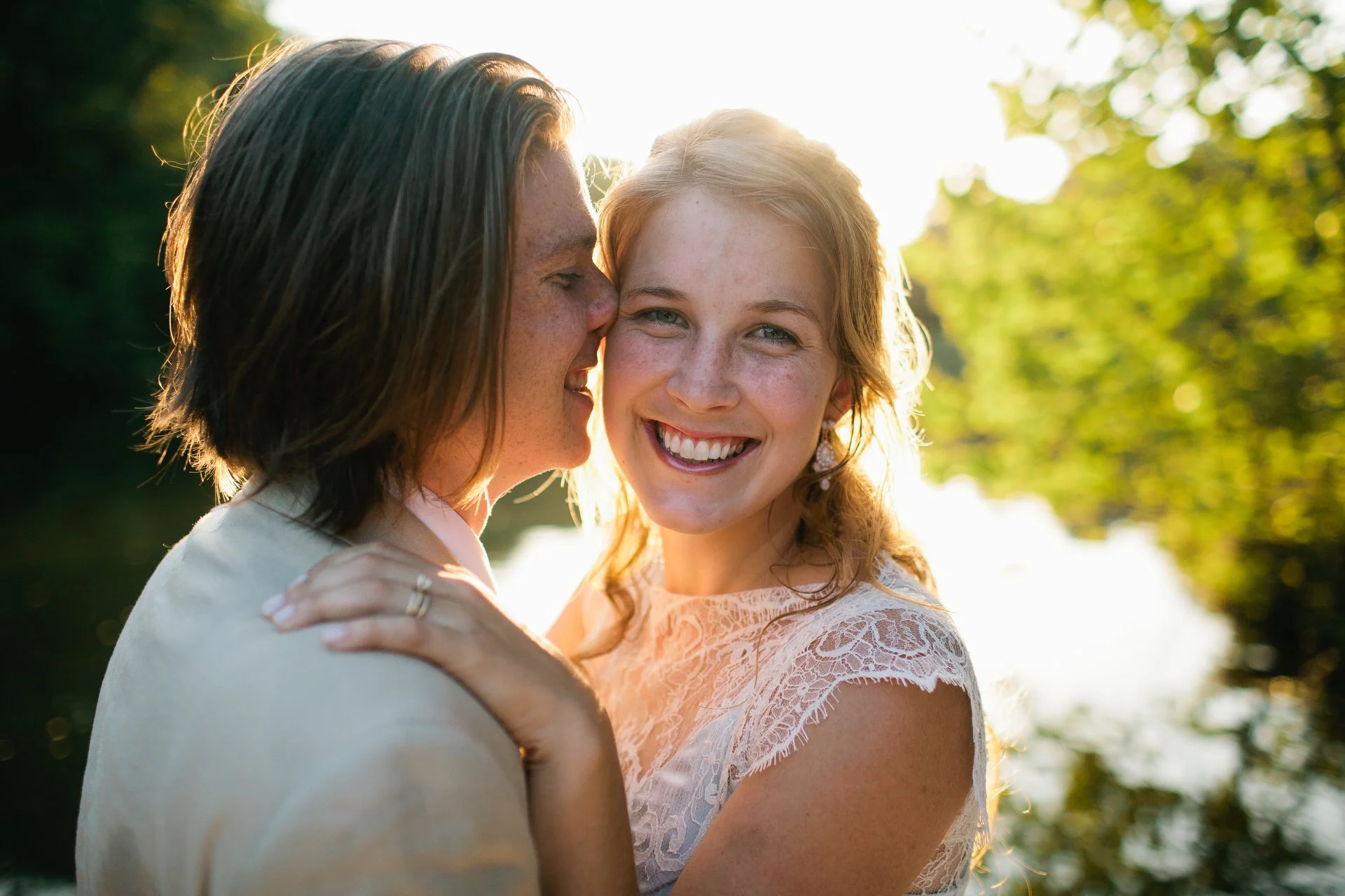 A woman with dark hair and a woman with light red hair smiling with their eyes closed outdoors during sunset, surrounded by green trees.