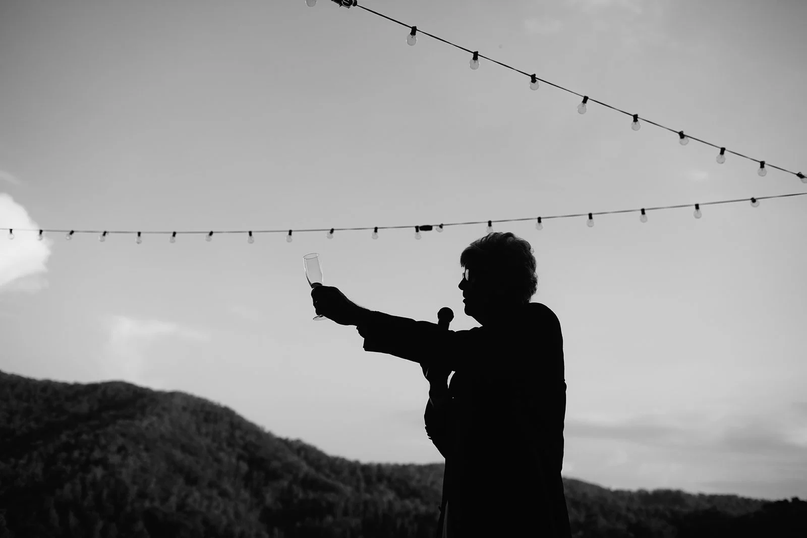 Silhouette of a woman with glasses holding a champagne glass and a microphone, raising her arm, outdoors with string lights overhead and hills in the background.