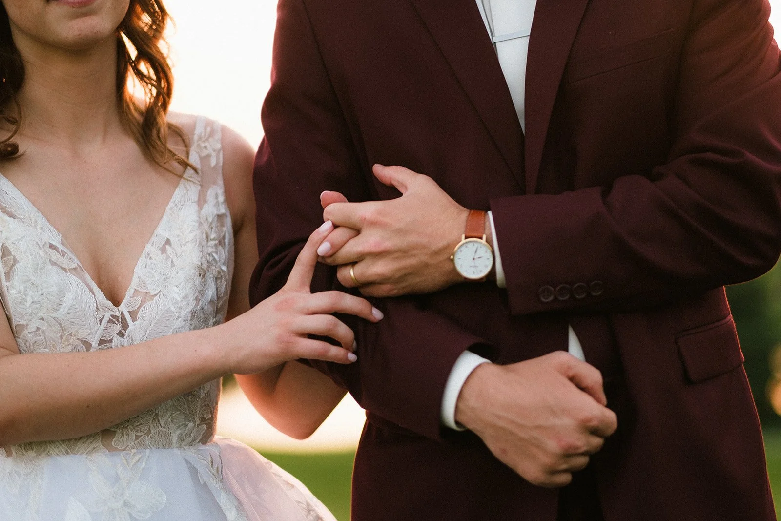 A woman holding a man's hand during a wedding ceremony, with both dressed in formal attire. The woman wears a white lace dress, and the man wears a burgundy suit with a watch on his wrist.