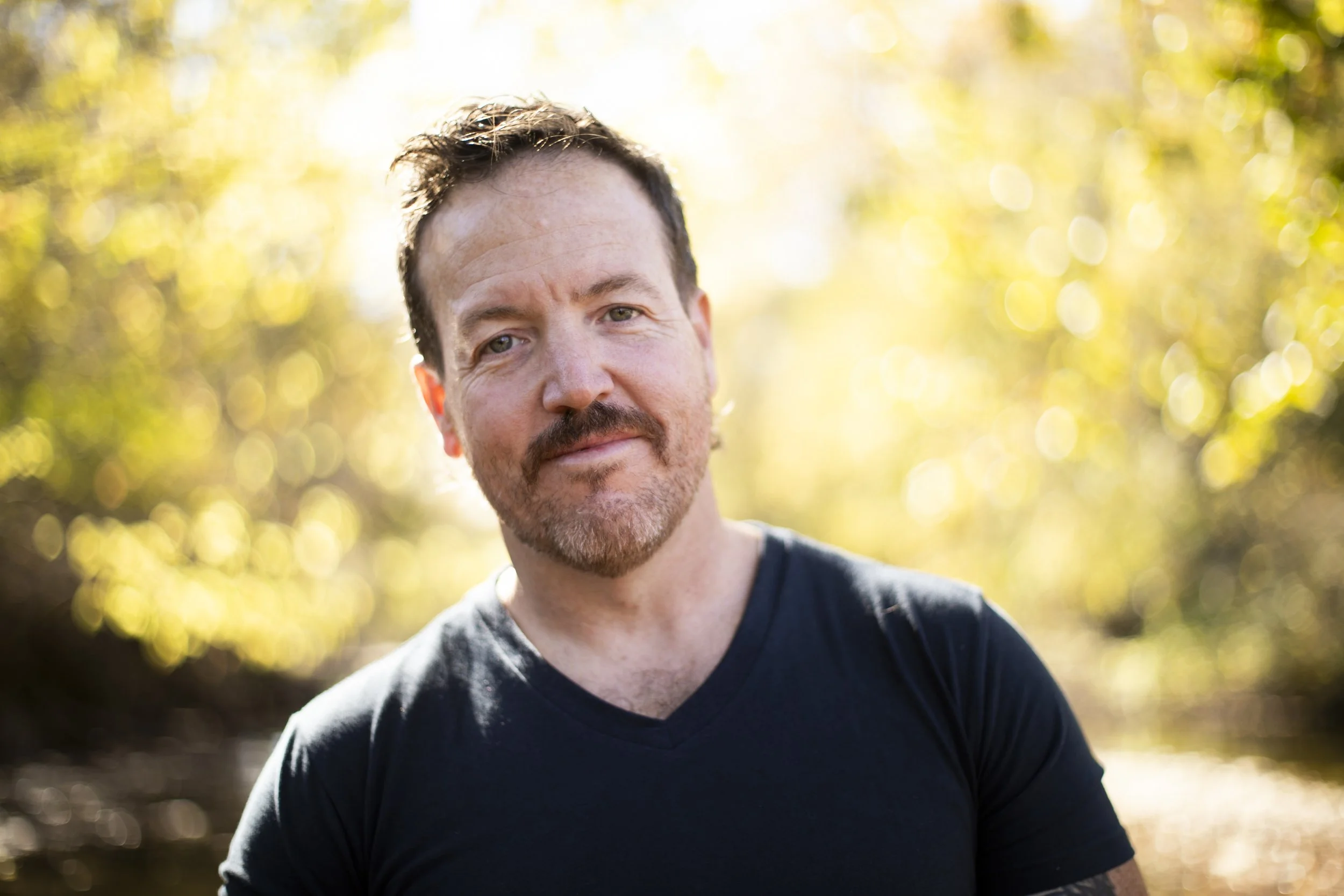 A man with short dark hair, a beard, and mustache smiling outdoors with sunlight filtering through yellow-green trees in the background.