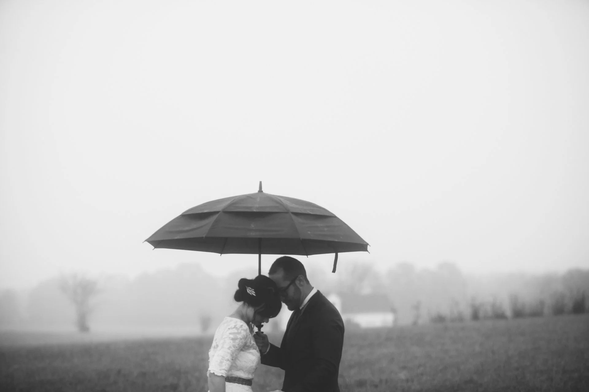 Black and white photo of a couple under an umbrella during rainy weather, close together, with the man's forehead touching the woman's.