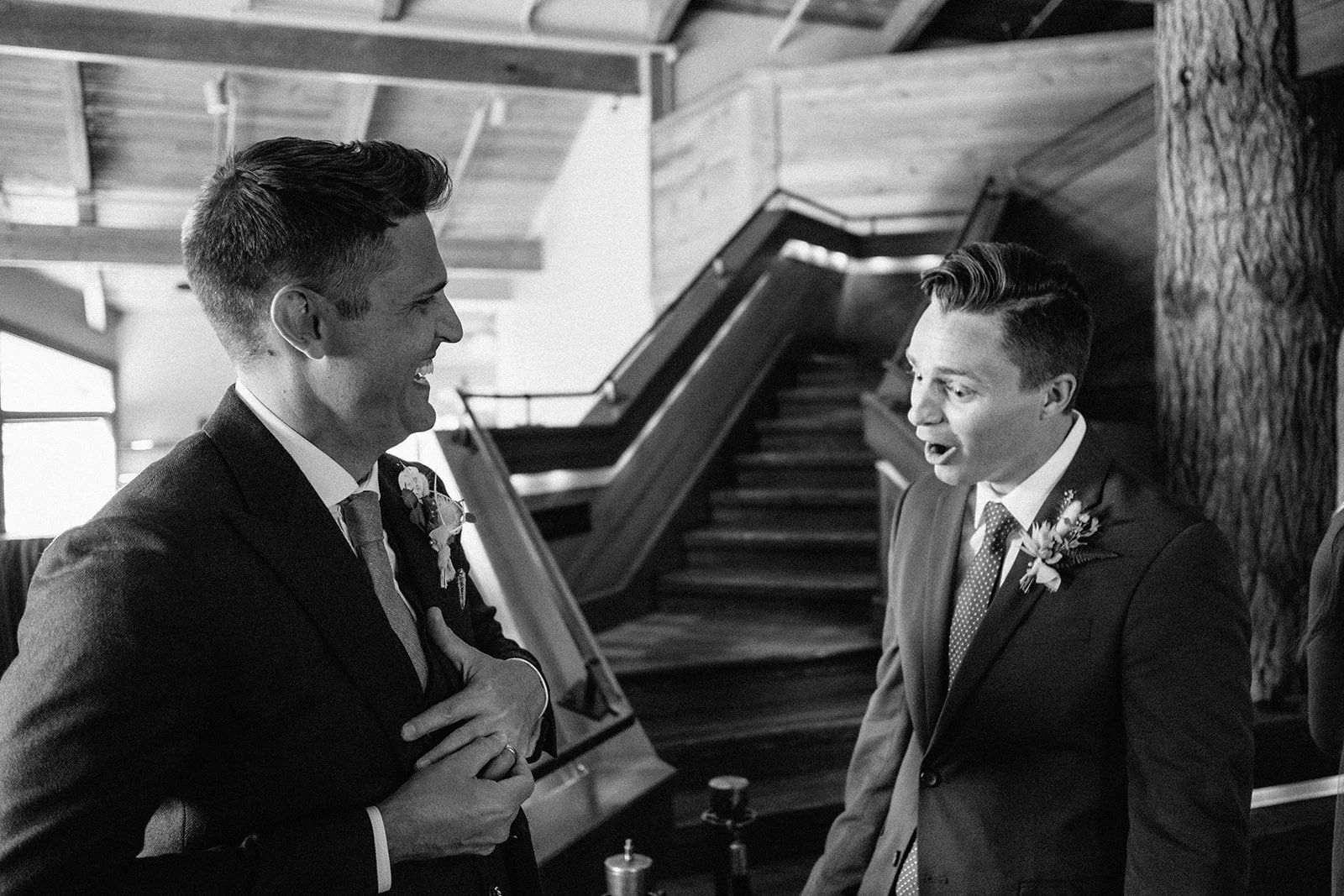 Two men in suits with boutonnières, smiling and talking indoors near staircase and a large tree trunk, in black and white.