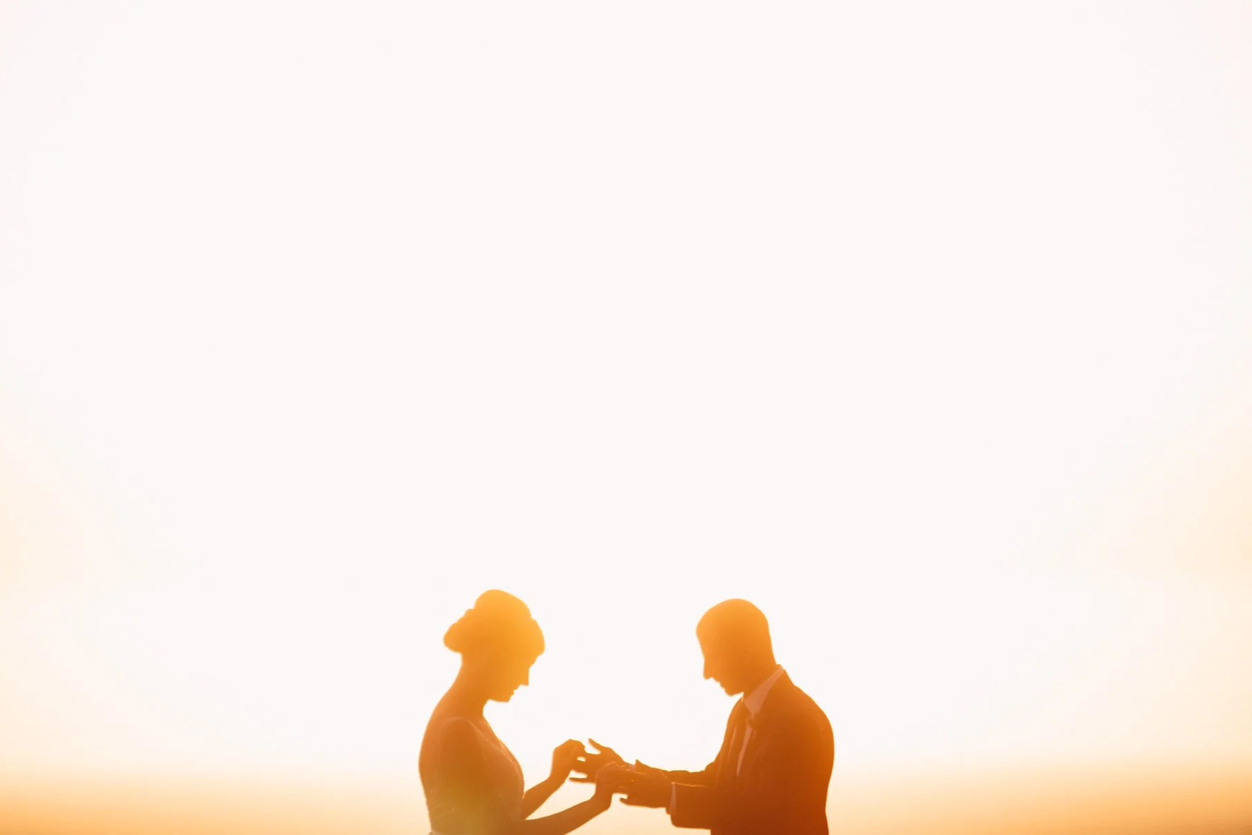 Two people facing each other and engaging in a hand gesture during sunset, with their silhouettes visible against the bright sky.