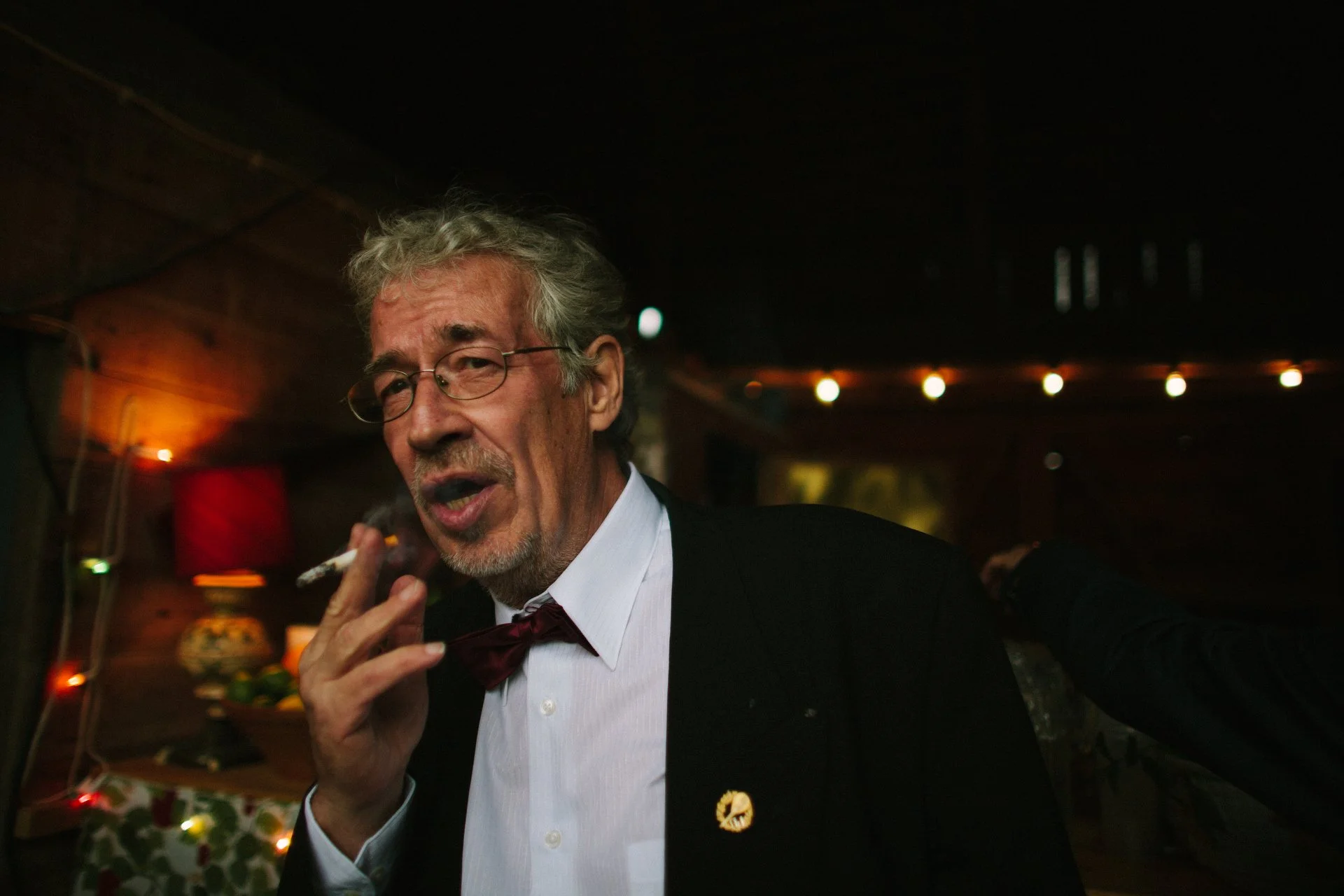 An elderly man in a tuxedo with a red bow tie, glasses, and gray hair, smoking a cigarette in a dimly lit room with festive holiday decorations and warm lighting.