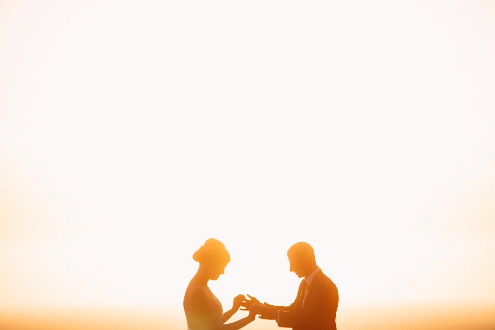 Silhouettes of a man and woman facing each other, holding hands, against a sunset background.