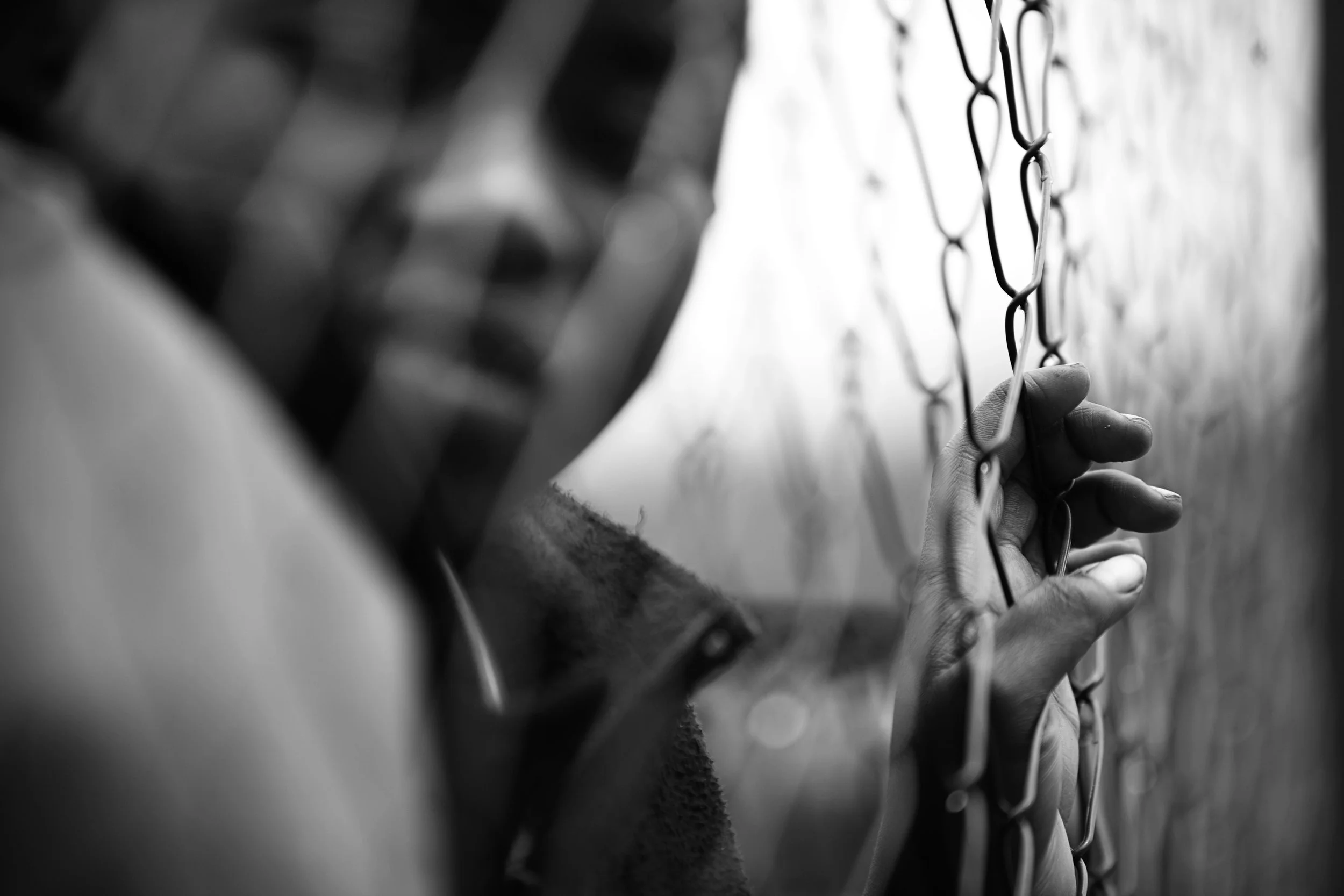 Black and white photo of a person gripping a chain-link fence with their hand.
