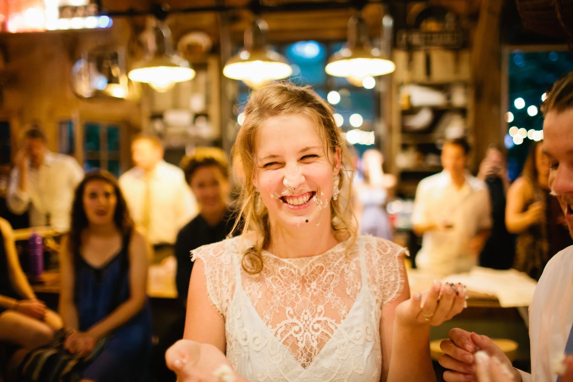 A woman with lace dress smiling with cake on her face surrounded by friends at a celebration.