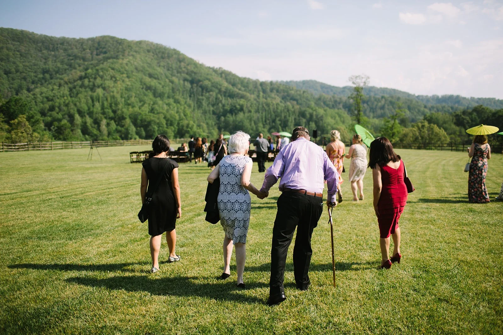 People attending an outdoor gathering or wedding on a grassy field with mountains in the background, some holding umbrellas.