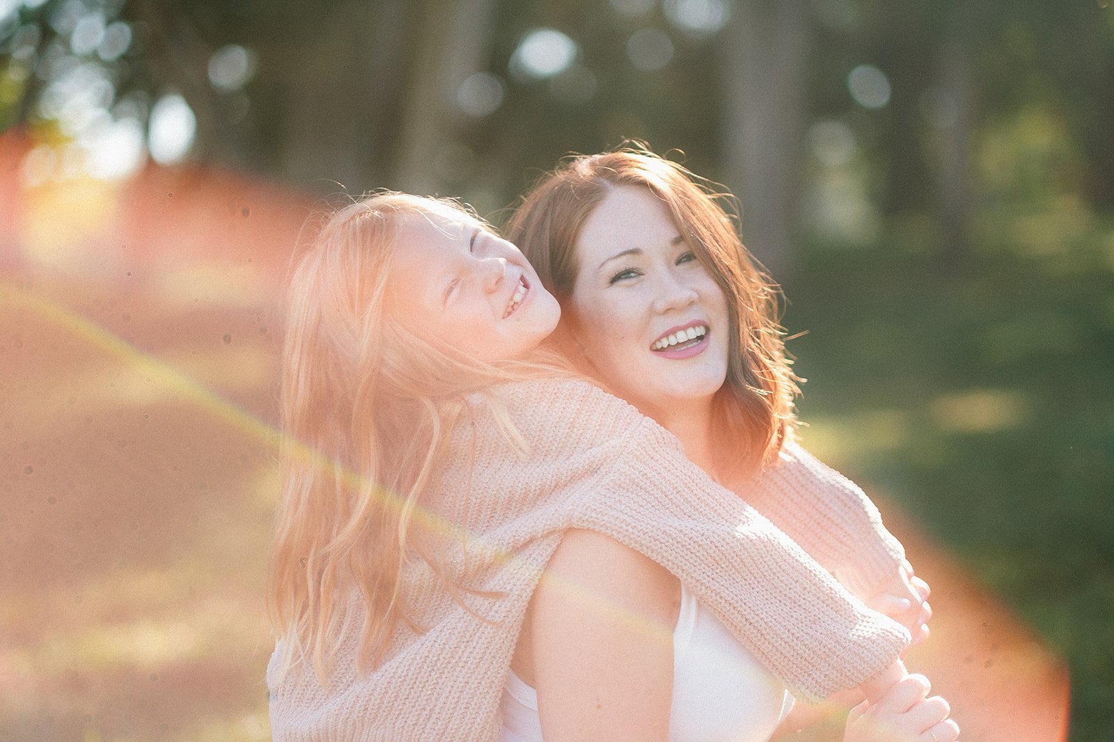 Two women are outdoors, smiling and enjoying a moment together. One is holding the other on her back, in a joyful and affectionate embrace, with sunlight creating a lens flare.