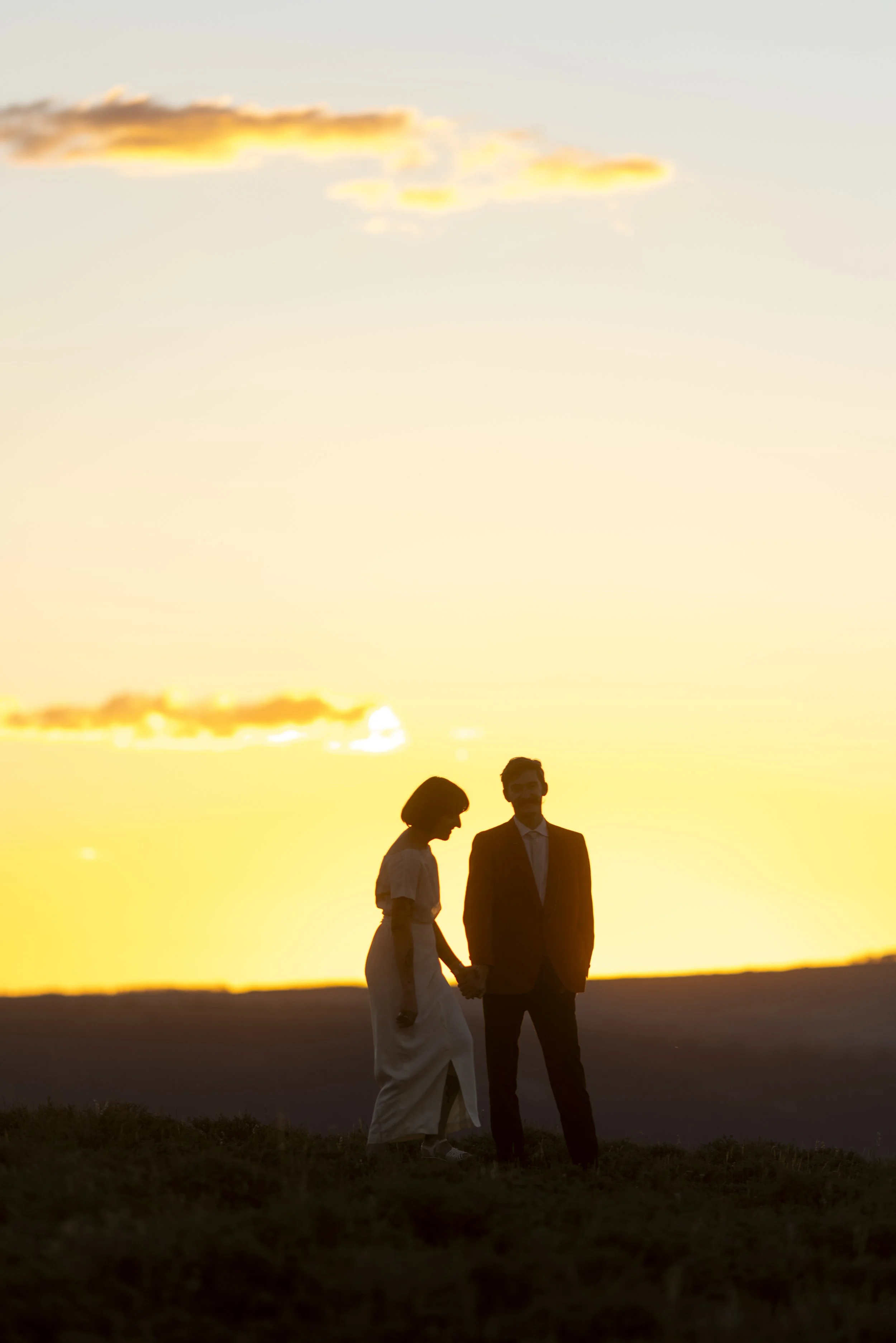 A couple holding hands and standing together outdoors during sunset, with the sky glowing with orange and yellow hues.
