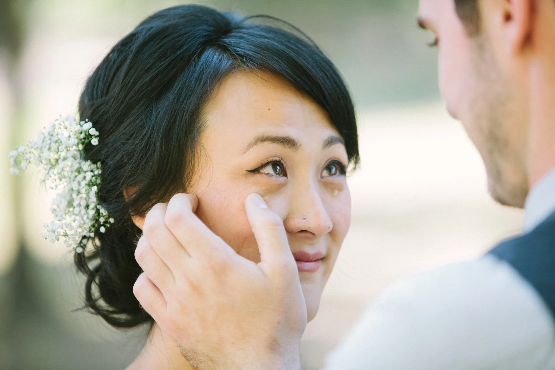A bride with black hair in an updo decorated with small white flowers, is having her makeup touched up by a person in a suit, with the person's hand gently pinching her cheek and adjusting her makeup.