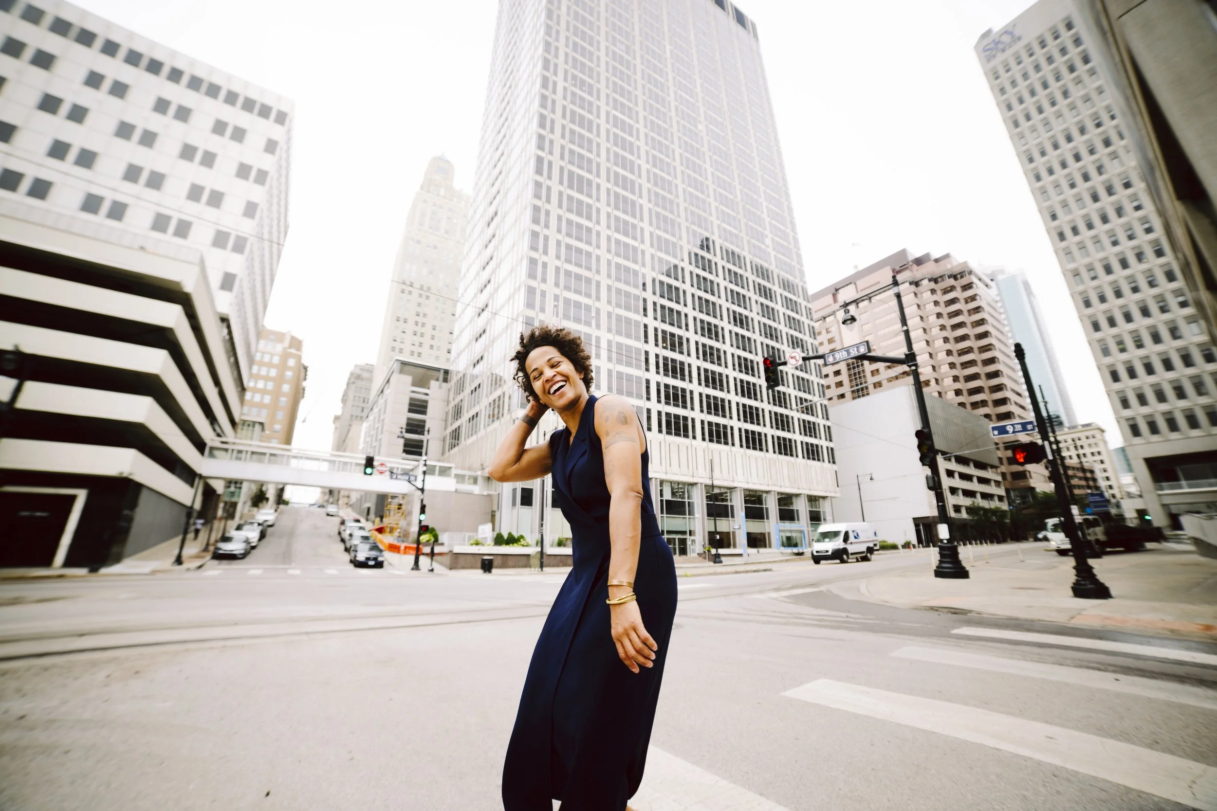 A woman in a long dark dress standing on a crosswalk in an urban city with tall buildings, smiling and touching her hair.