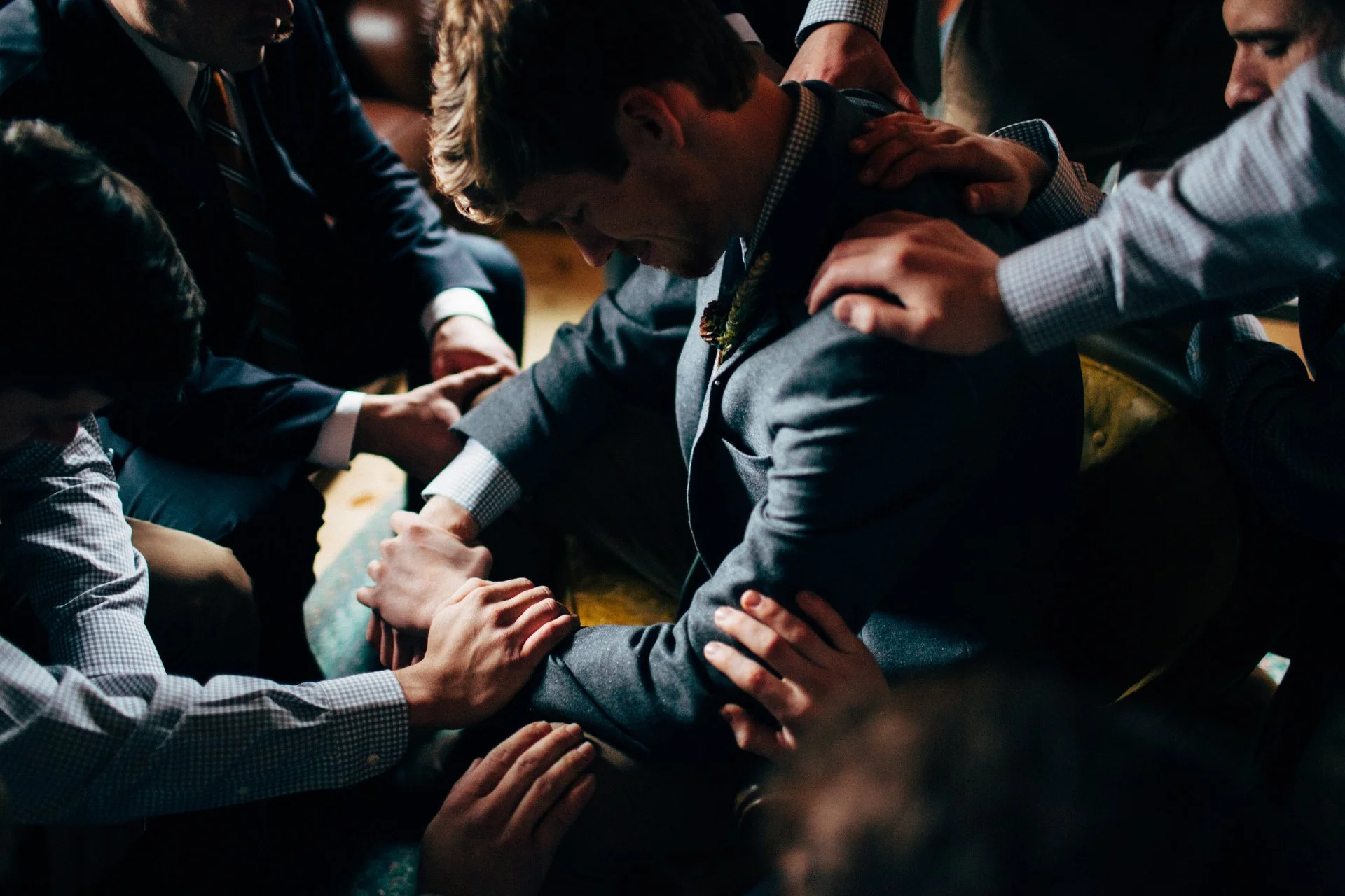 Group of men wearing formal suits, praying together with their hands clasped, and one man in the center with his head bowed and eyes closed.