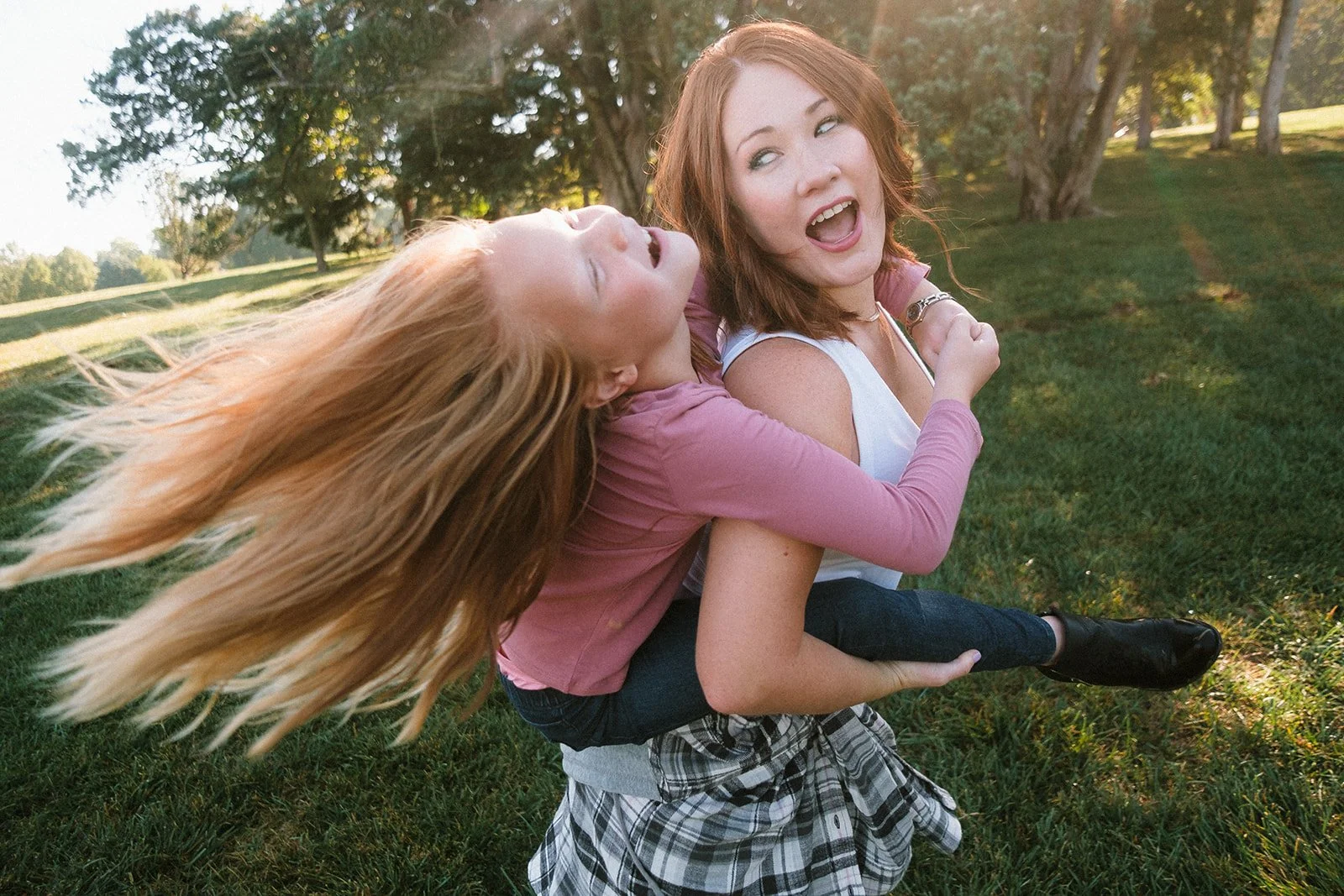 Two young women enjoying a playful moment outdoors on a sunny day, with one carrying the other on her back in a park with trees and grass.